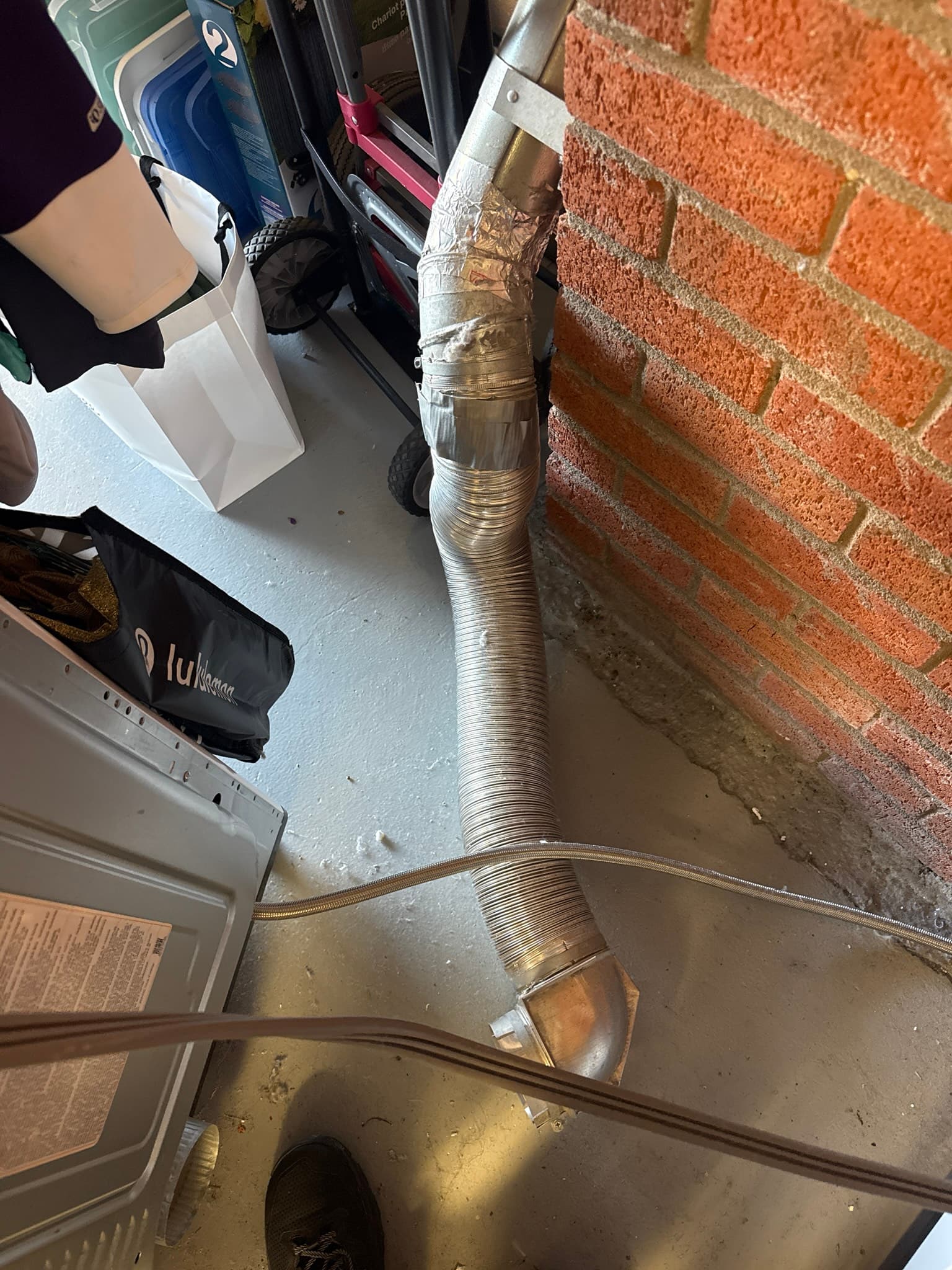 Flexible aluminum dryer exhaust duct wrapped with foil tape at a repair point, running along a brick exterior wall to a foundation penetration with a loose and inadequately sealed wall connection