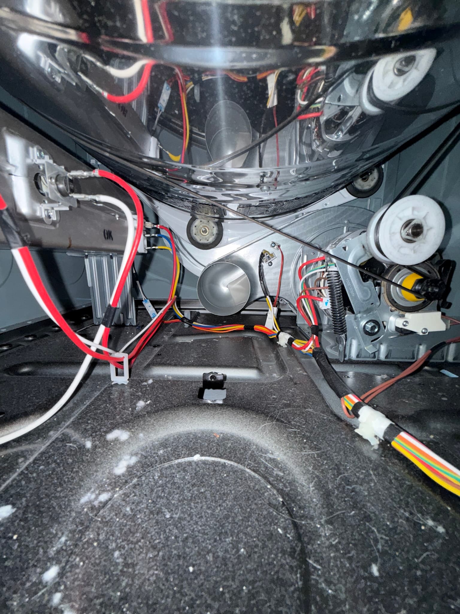 Interior view of a clothes dryer drum assembly with the panel removed, showing lint and dust accumulation on internal components including pulleys, thermostats, heating elements, and wiring harnesses.