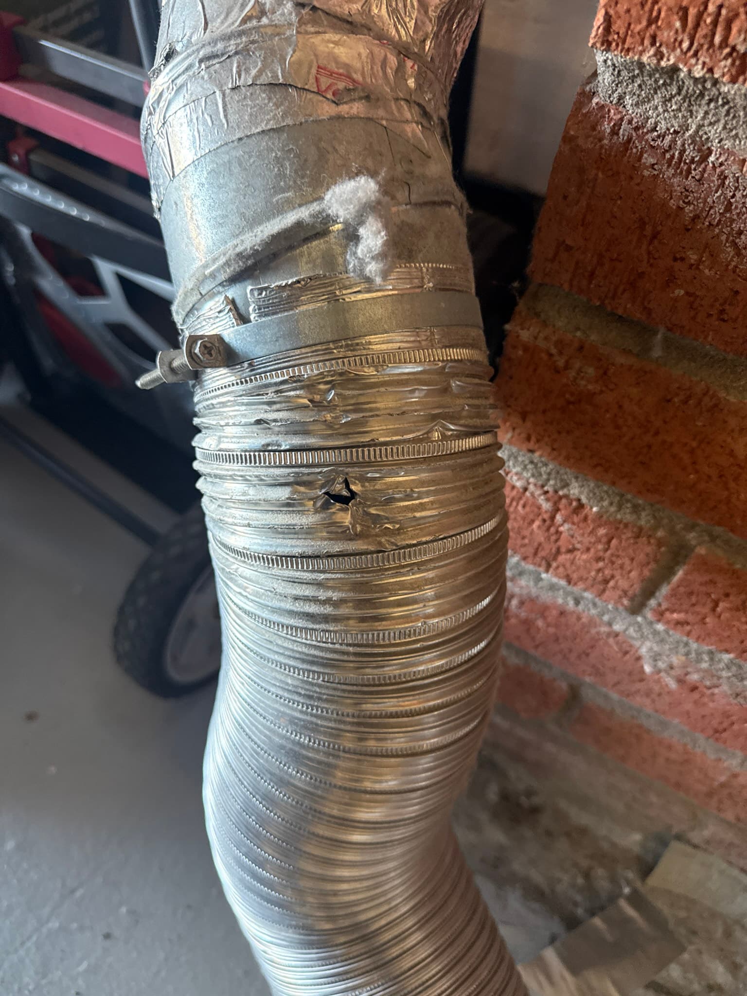 Flexible dryer vent duct secured with hose clamps near a brick wall, with heavy dust and lint buildup coating the duct exterior indicating restricted airflow and poor exhaust conditions