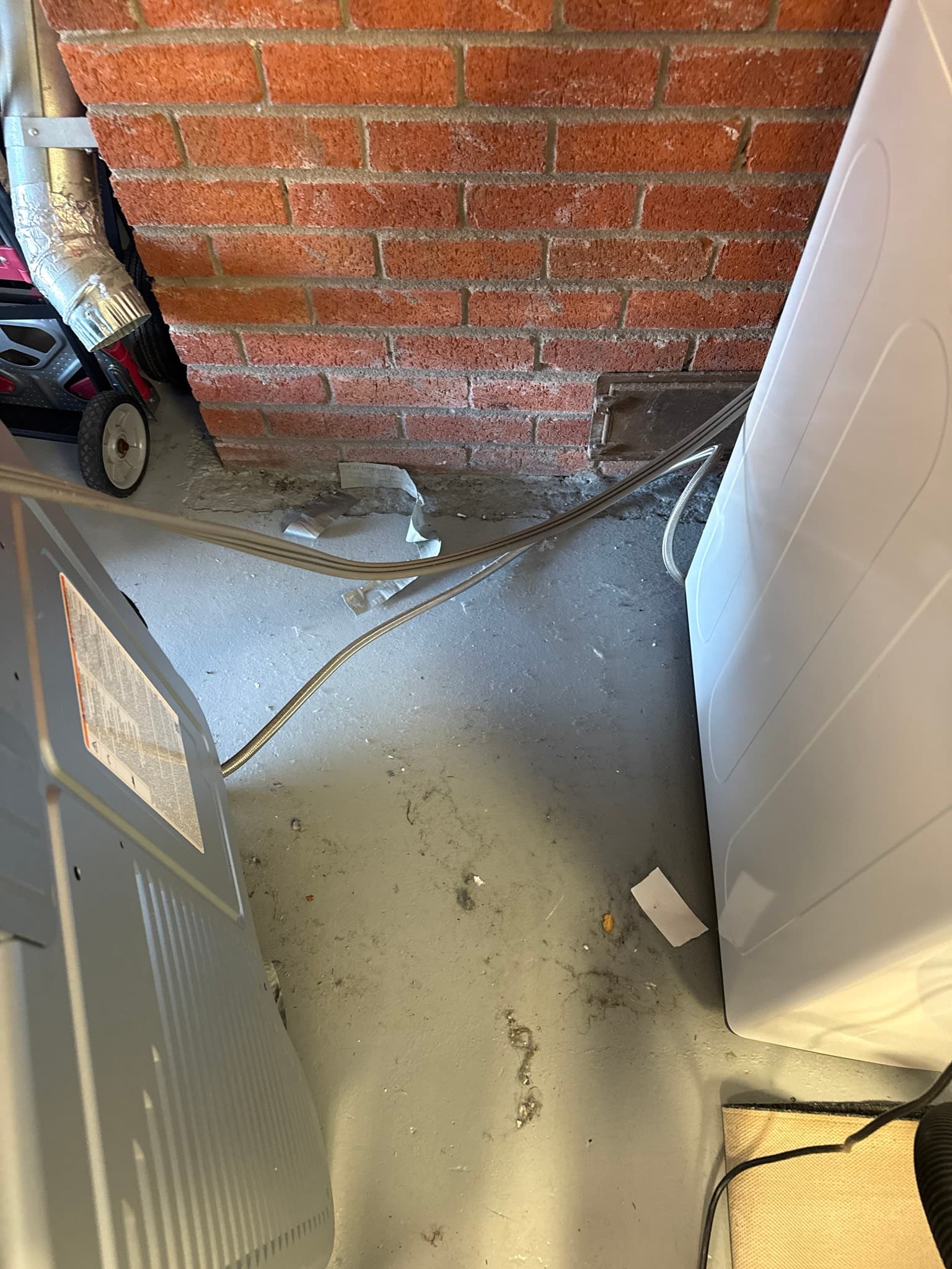 Dryer exhaust duct exiting the back of the dryer and running along a concrete floor to a rectangular penetration in a brick wall, with dust and debris visible on the floor around the unsupported duct