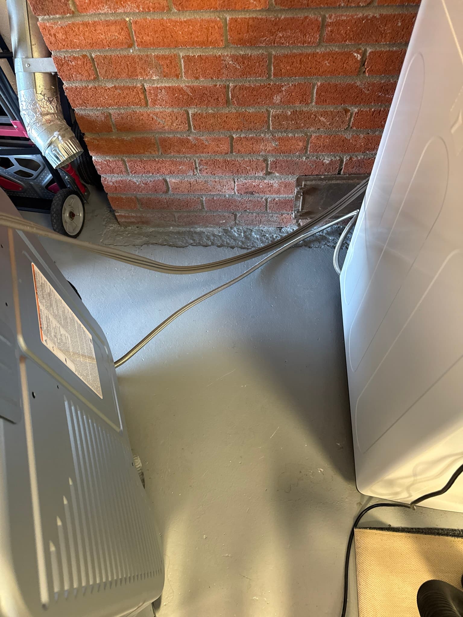 Metallic dryer exhaust duct connected to the rear of a clothes dryer against a brick wall in a garage, showing dust accumulation on the duct exterior prior to service