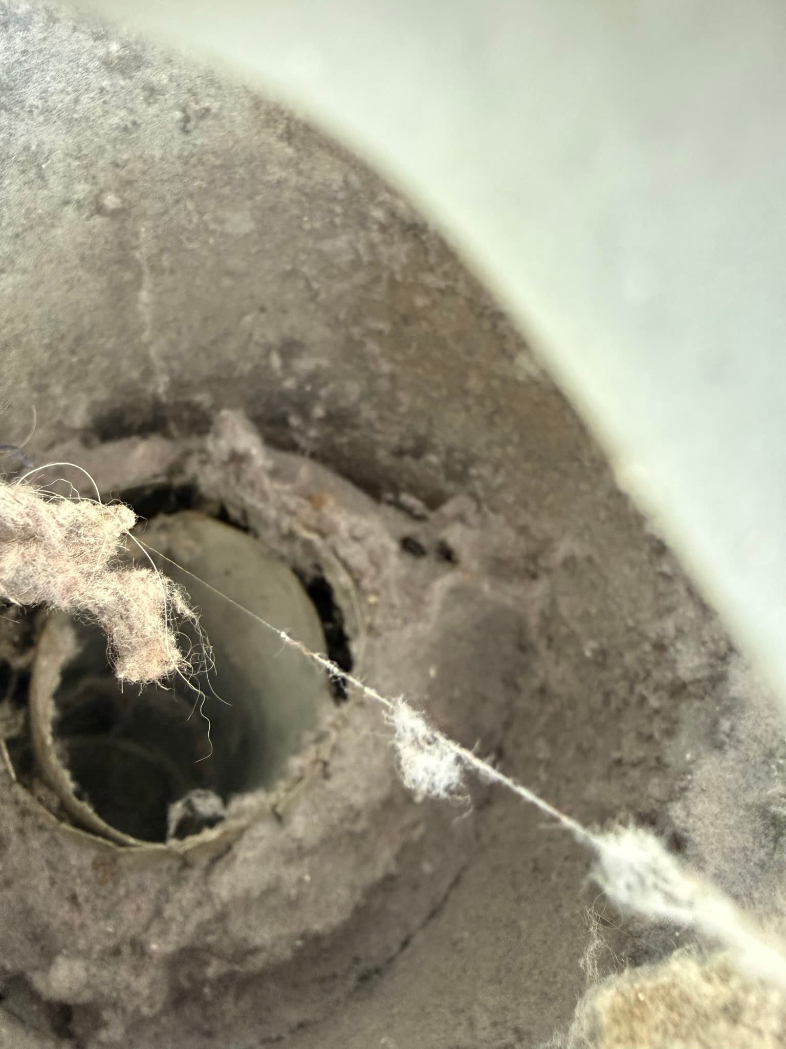 Exterior dryer vent cap opening heavily clogged with a dense mass of compressed lint fibers, with additional lint strands visible around the perimeter of the metal periscope-style cap.