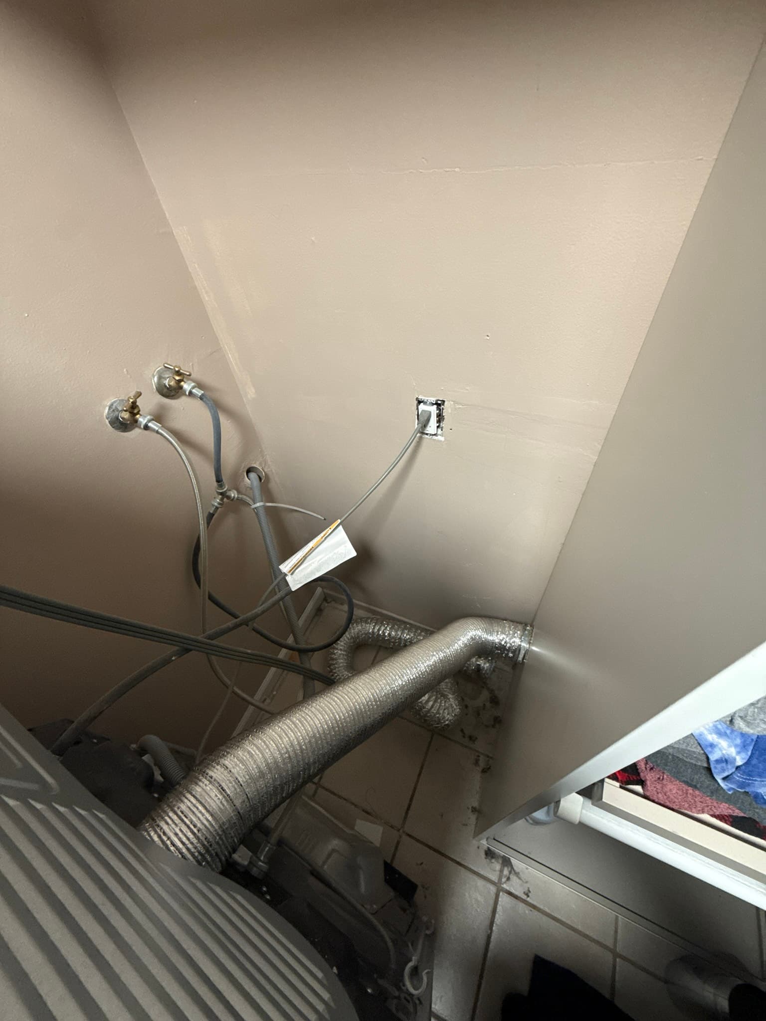 A metallic flexible dryer vent duct running from a dryer unit toward an exterior wall outlet, with water supply lines mounted on the wall above and an electrical junction box visible nearby.