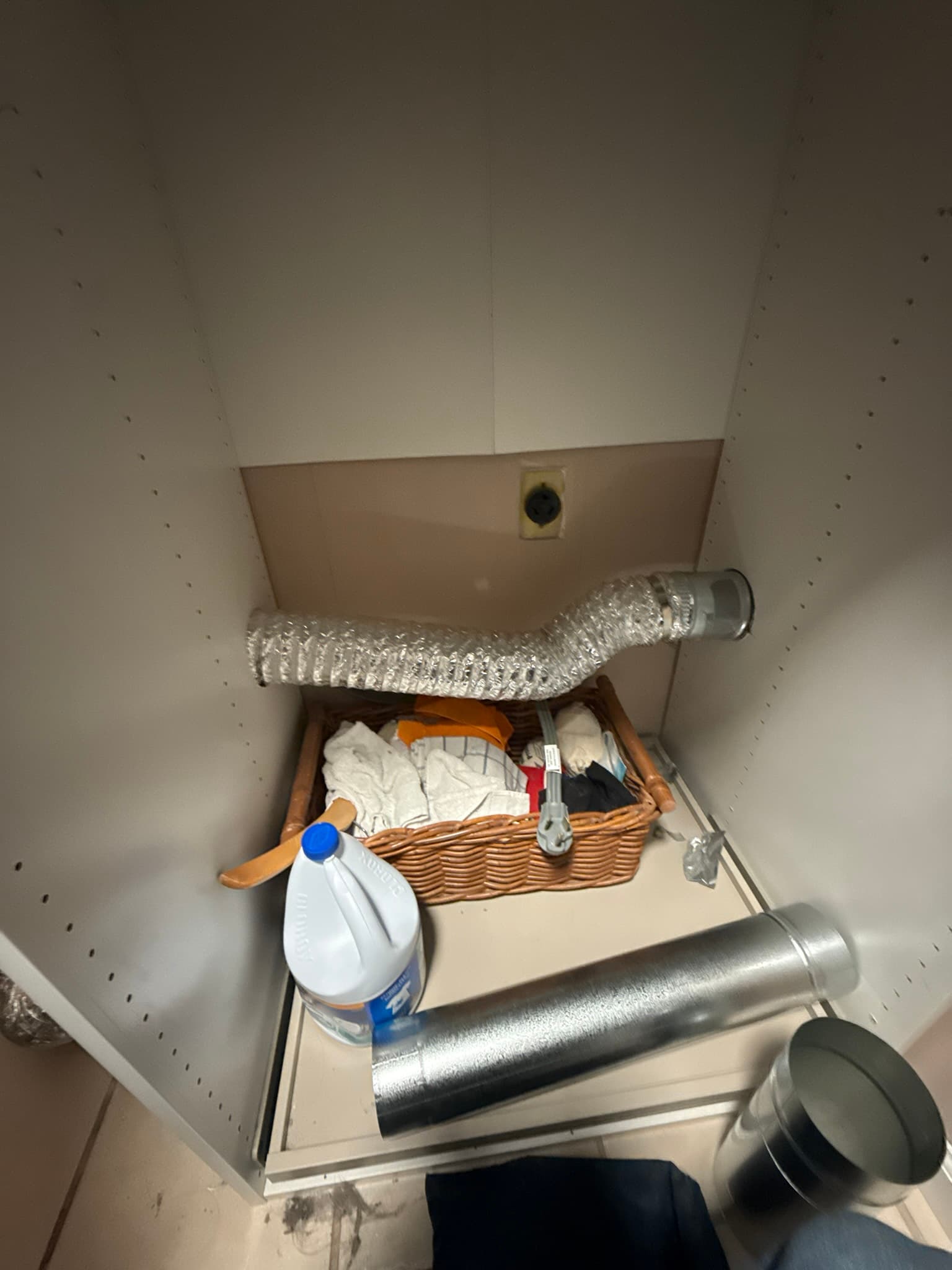 Interior of a laundry closet showing a clean flexible aluminum dryer vent duct running horizontally, with rigid metal duct components, elbows, and tools stored in a basket below, and a round wall penetration visible at the upper rear.