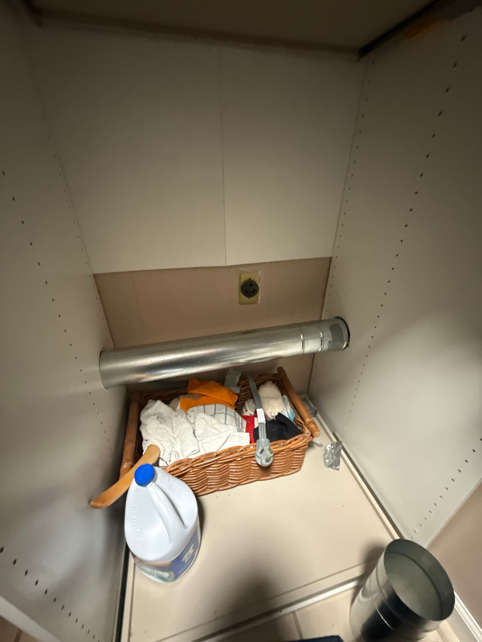 Interior of a storage shelf with a rigid metal dryer vent duct installed horizontally, a beige electrical outlet on the back wall above the duct, and household items including a wicker basket, folded linens, and a handheld fan stored on the shelf.