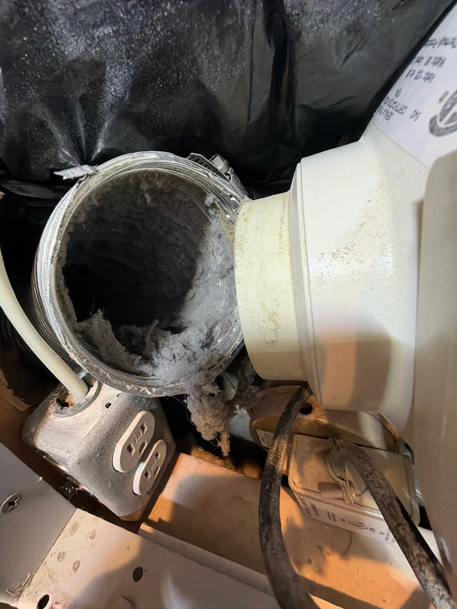 Close-up of the dryer's rear transition duct connection heavily coated with lint, with loose clumps spilling out near an electrical outlet