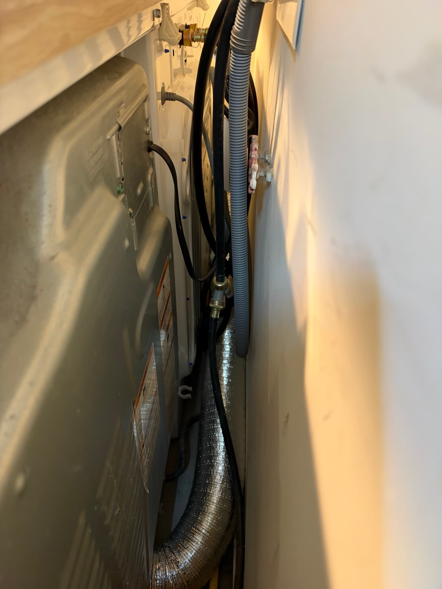 Tight gap behind a dryer showing the foil transition duct, water supply lines, and electrical cord feeding into the wall opening near the booster fan