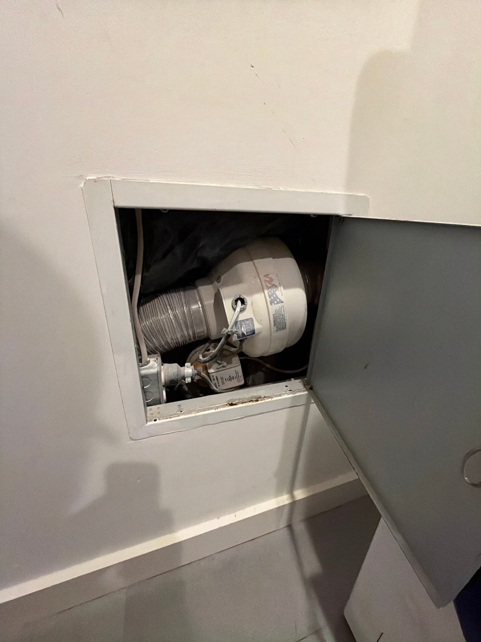 Access panel exposing an inline booster fan connected to foil transition duct in a recessed wall cavity, with wiring and a junction box visible alongside heavily lint-saturated ductwork
