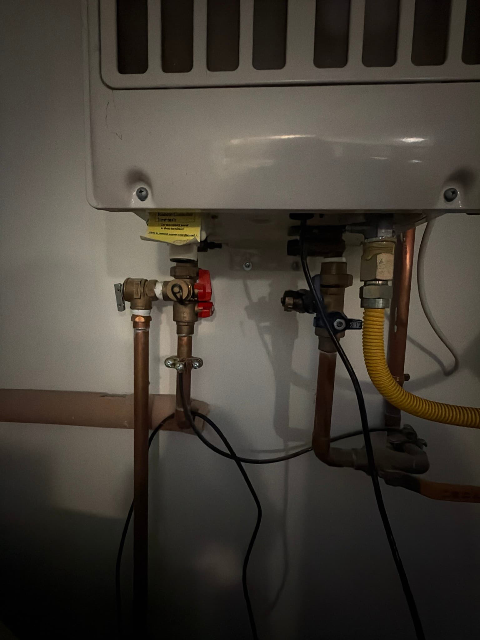 Underside connections of a tankless water heater showing copper water lines, red and black handled isolation valves, and a corrugated yellow gas connector
