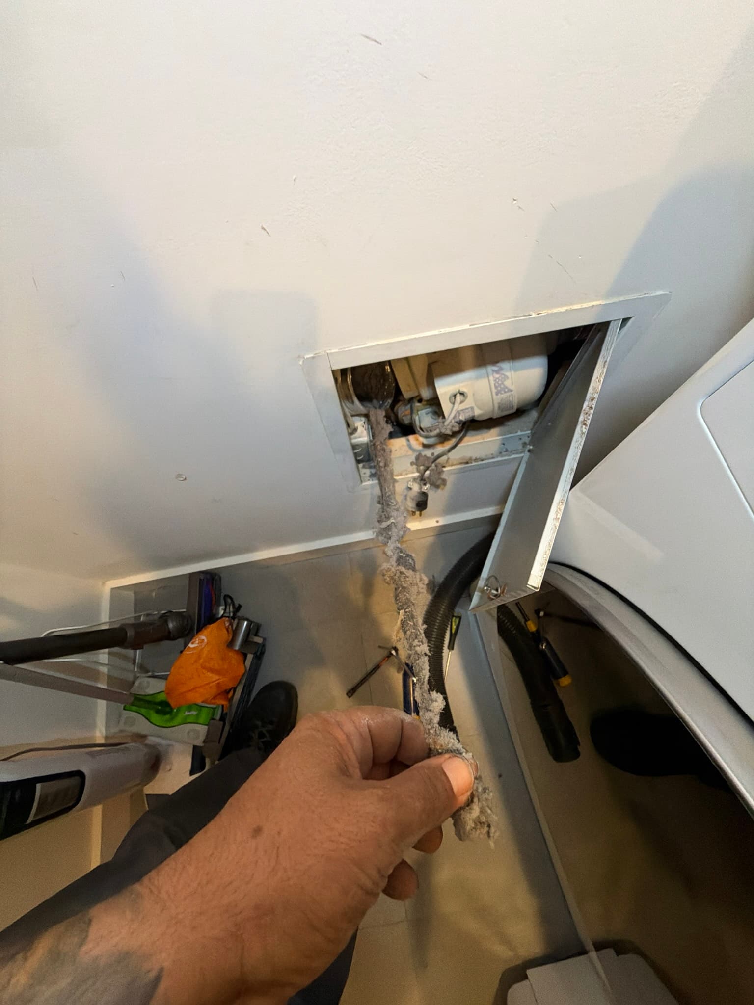 Open access panel behind a dryer showing semi-rigid metal duct heavily coated in matted lint, with a gloved hand pulling a compacted lint clump near a booster fan and electrical outlet in a tight wall cavity