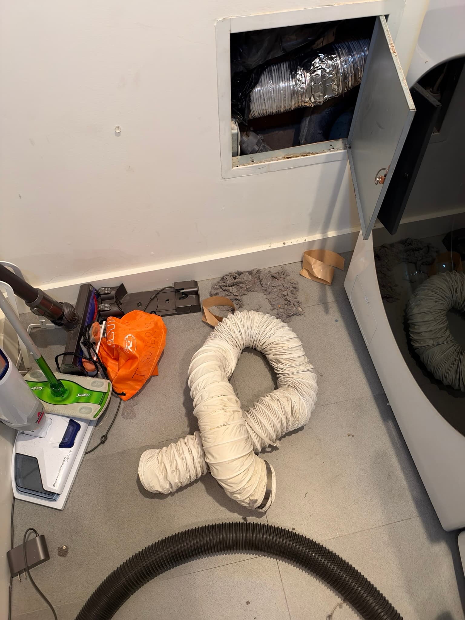 Wall access panel opened to show the exhaust duct cavity with a disconnected white flexible transition duct and a substantial pile of extracted lint and debris on the floor
