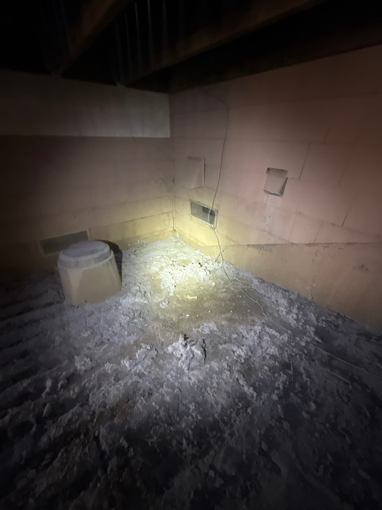 Attic space with heavy lint accumulation covering the floor and surrounding surfaces, with a white bucket, electrical wiring, and rectangular metal duct openings visible, indicating a dryer exhaust system failure that allowed lint to escape into the attic