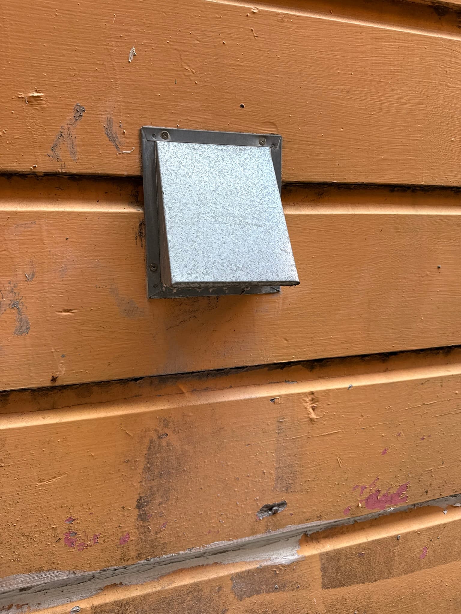 Exterior dryer vent cap mounted on wood siding showing a metal frame with a white damper flap. The damper surface has light dust and lint deposits. Surrounding siding shows weathering and staining.