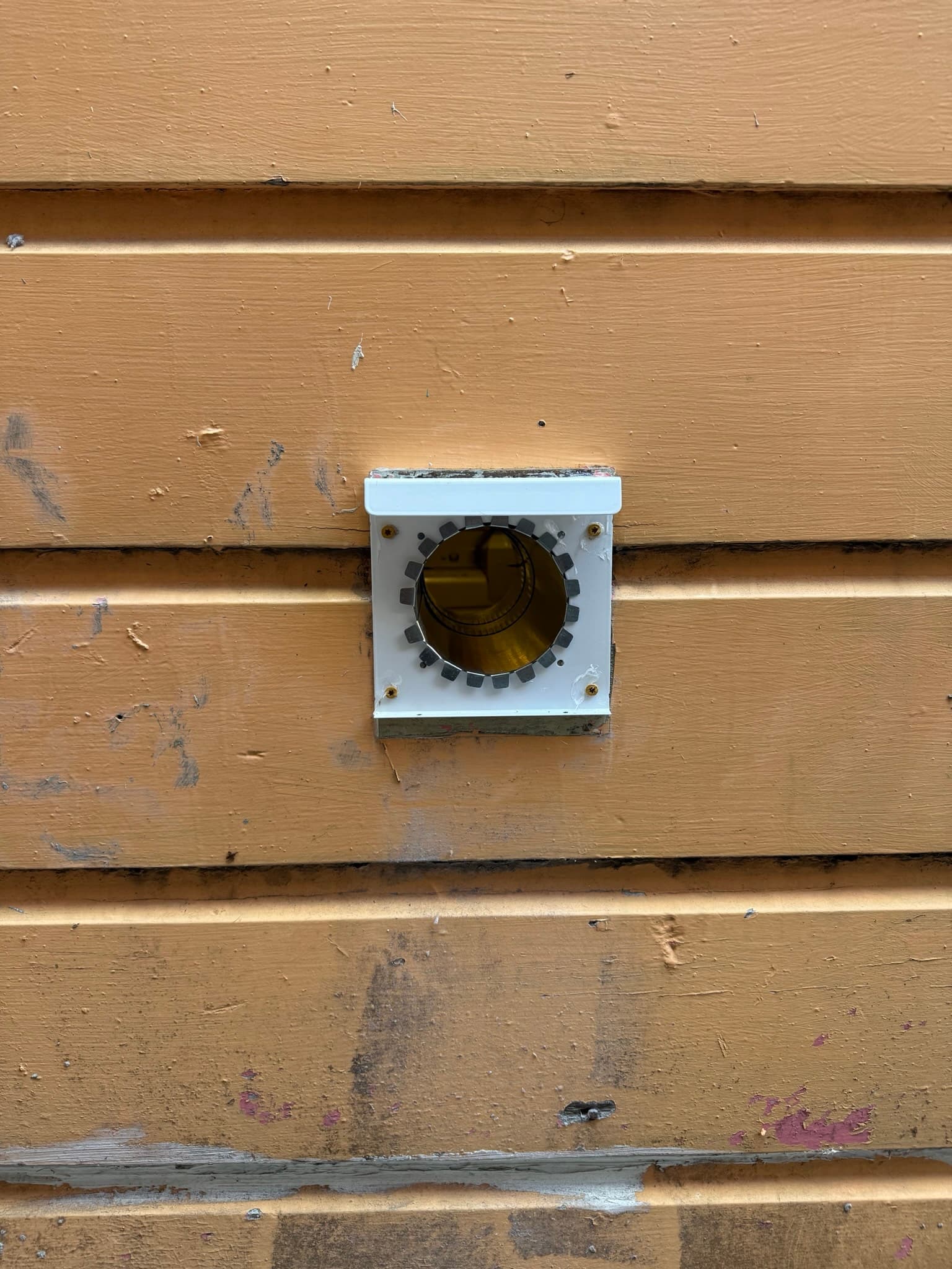 Exterior dryer vent cap on wood siding with a white trim ring, round damper opening, and visible flapper mechanism inside. Fasteners are visible on the trim ring. Surrounding siding shows weathering and paint deterioration.