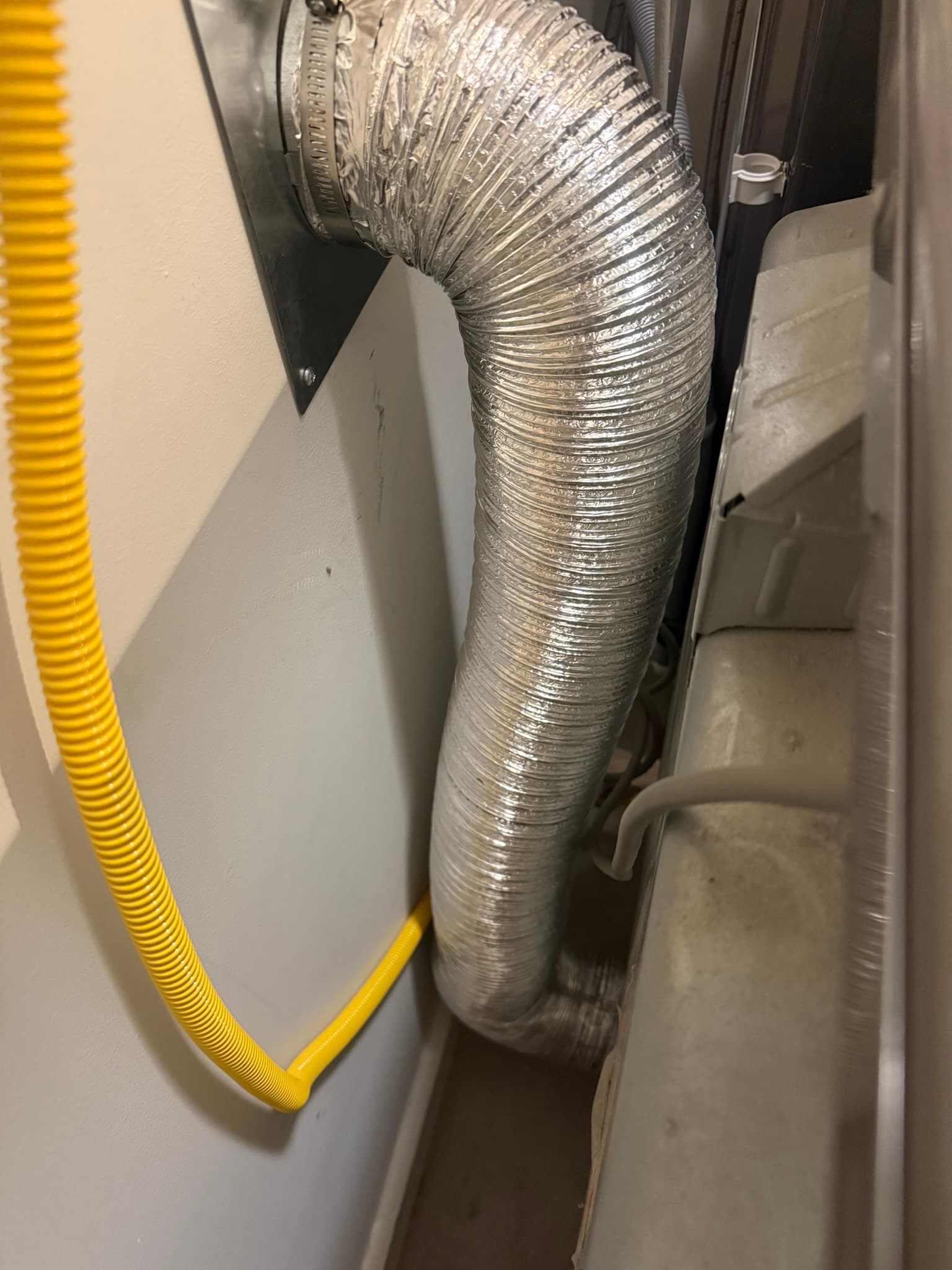 Rear of a dryer appliance showing a flexible aluminum transition duct connected to the dryer back panel with a hose clamp. The duct appears clean and properly connected, with no visible lint accumulation. A yellow flexible conduit is visible on the left side.