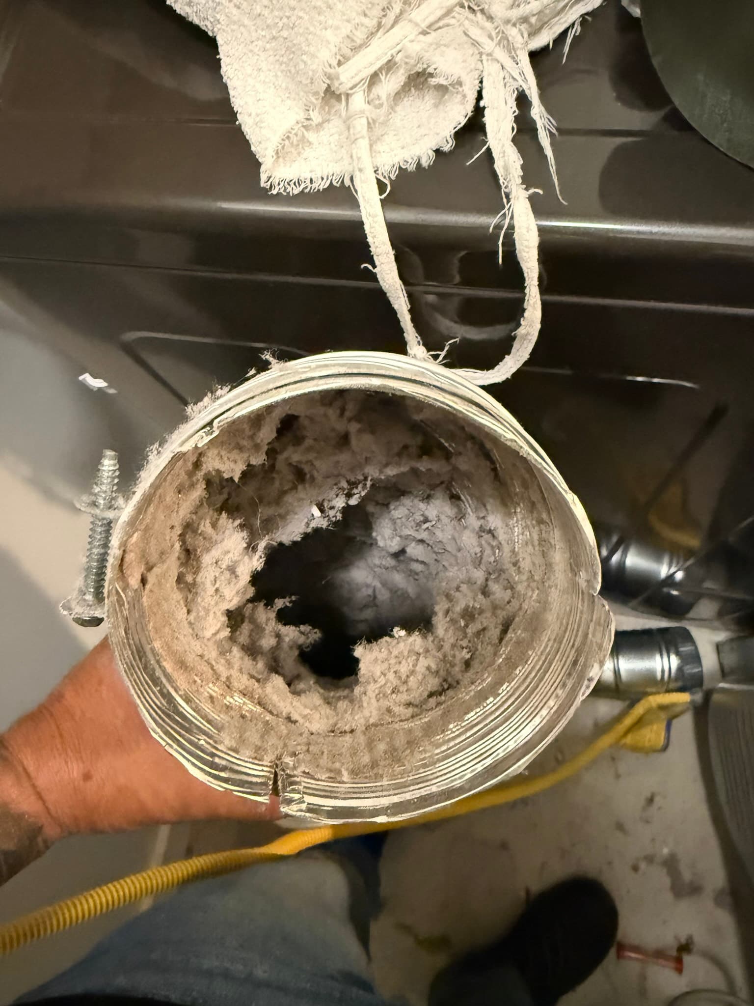 Interior view of a 4-inch metal dryer vent duct showing severe lint accumulation coating the walls, with a thick compacted lint plug visible at the center of the passage. Fabric suspension straps are attached to the duct exterior.