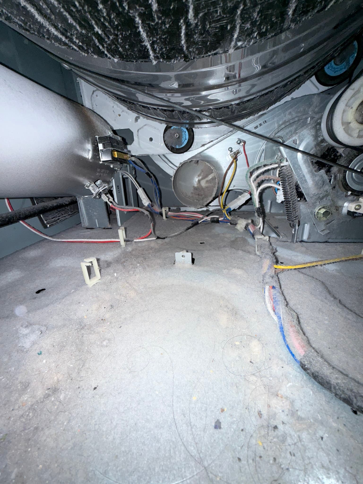 Interior underside view of a dryer drum and blower assembly showing substantial lint accumulation on the drum surface, blower wheel housing, and surrounding components including the thermal cutoff, idler pulley, and motor. Color-coded electrical wiring is visible.