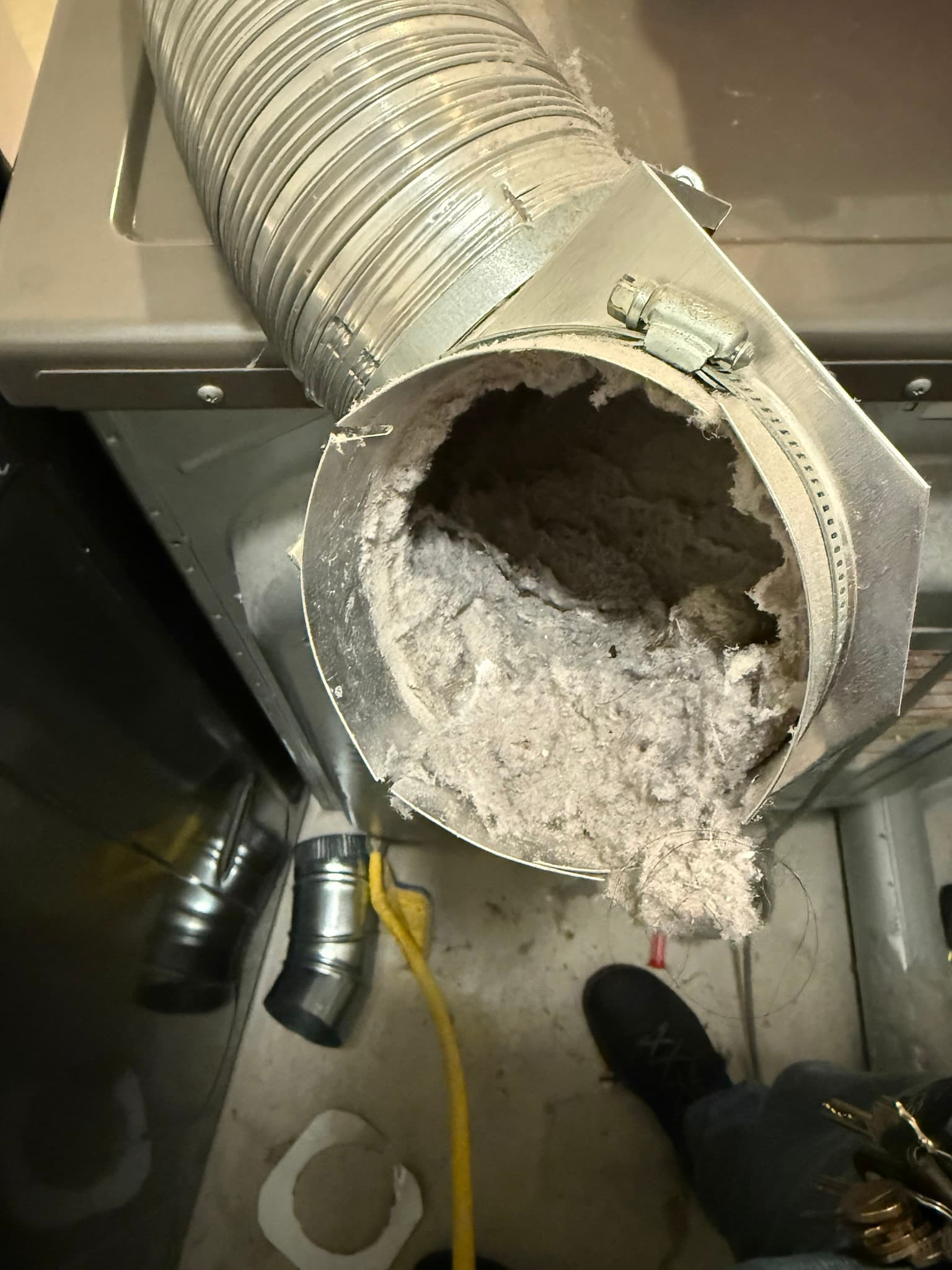 Severely lint-clogged flexible aluminum dryer vent duct connected to a transition box with hose clamps. The duct interior is nearly completely blocked by a dense compacted mass of lint, leaving almost no open airflow passage.