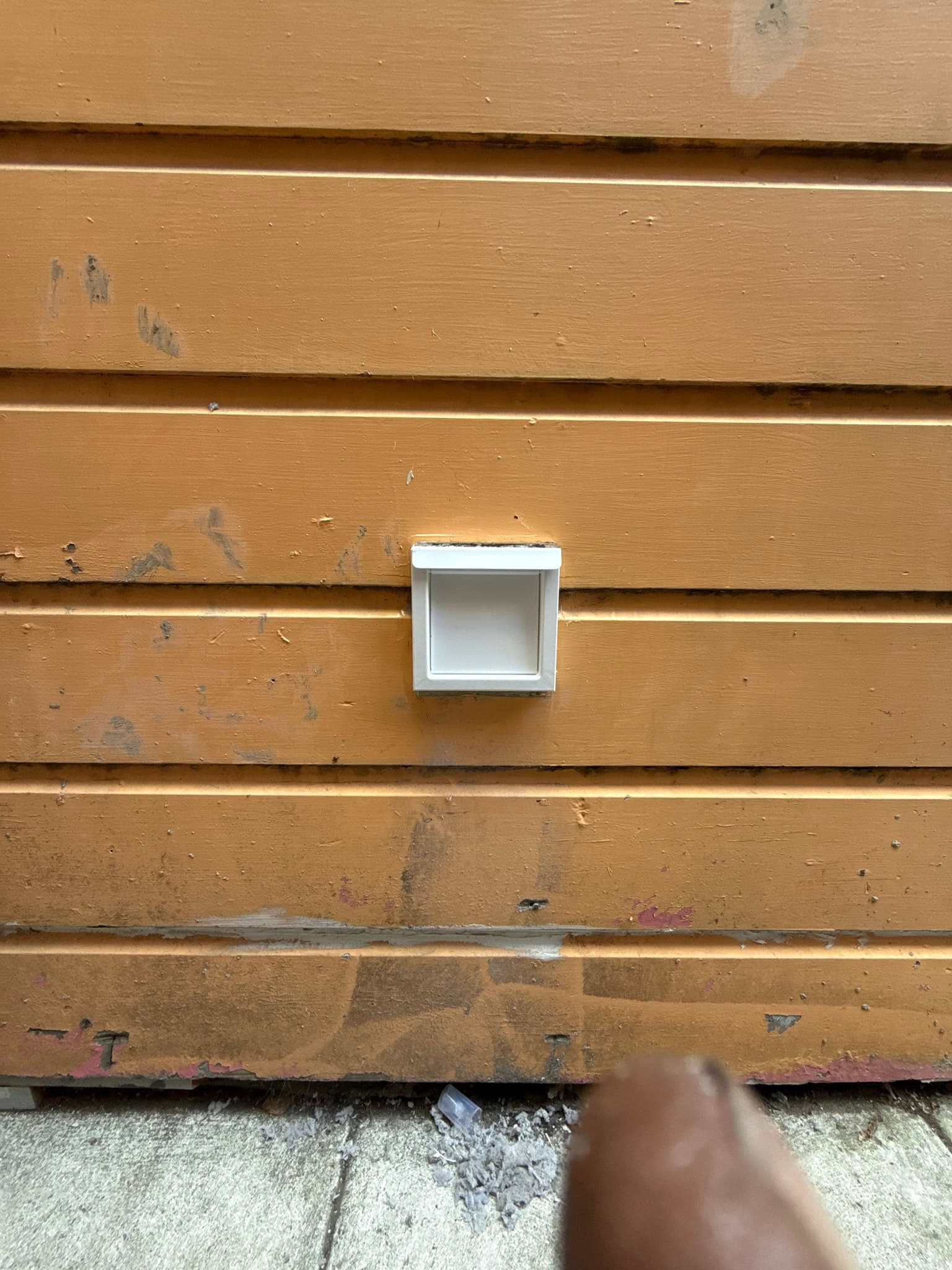 Exterior white periscope-style dryer vent cap mounted on tan horizontal lap siding. The vent opening appears clean and unobstructed. Surrounding siding shows weathering and surface dirt.