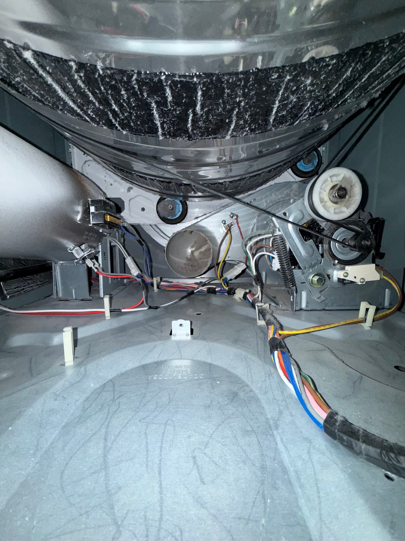 Interior view of a clothes dryer drum and mechanical assembly with severe lint accumulation coating the entire drum surface. The blower wheel, motor, idler pulley, and electrical connectors with colored wiring are visible and accessible beneath the heavy lint layer.