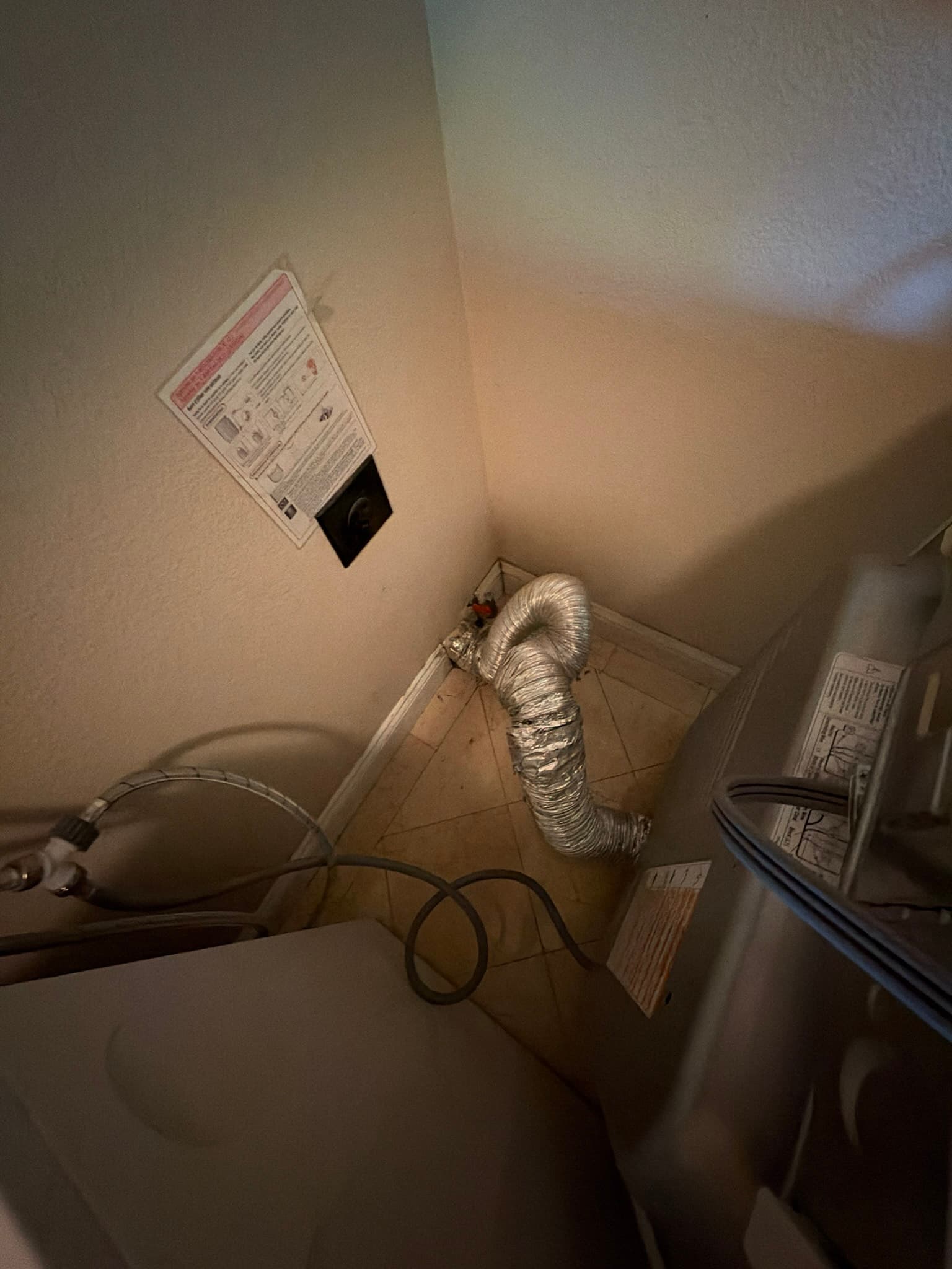 Foil flexible transition duct sagging with excess slack and doubling back sharply in the corner behind a dryer, next to a manufacturer warning placard on the wall.