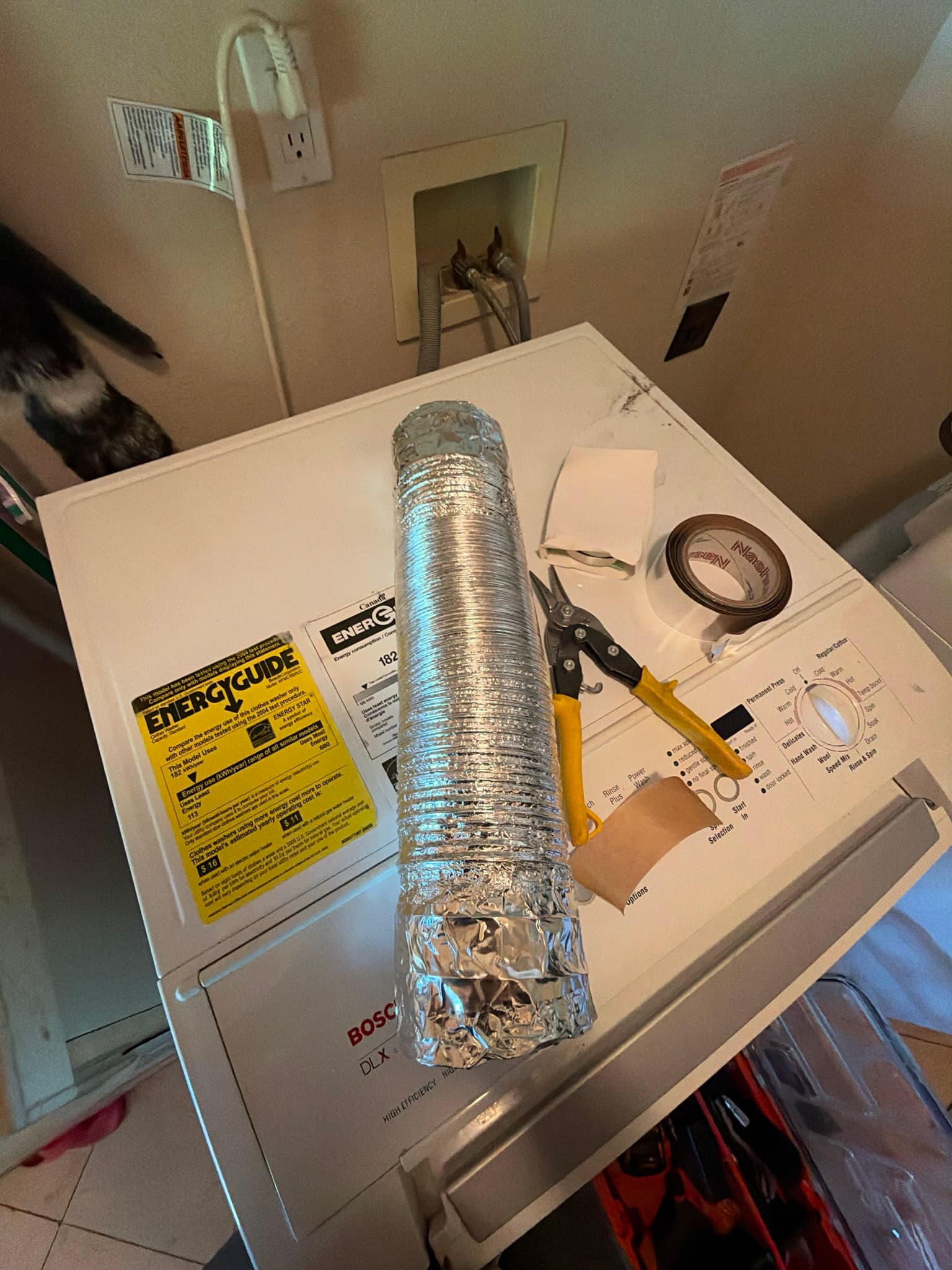 New section of DryerFlex transition duct resting on top of a Bosch dryer alongside tin snips, foil tape, and cloth, with the wall exhaust connection point visible and clear of debris.