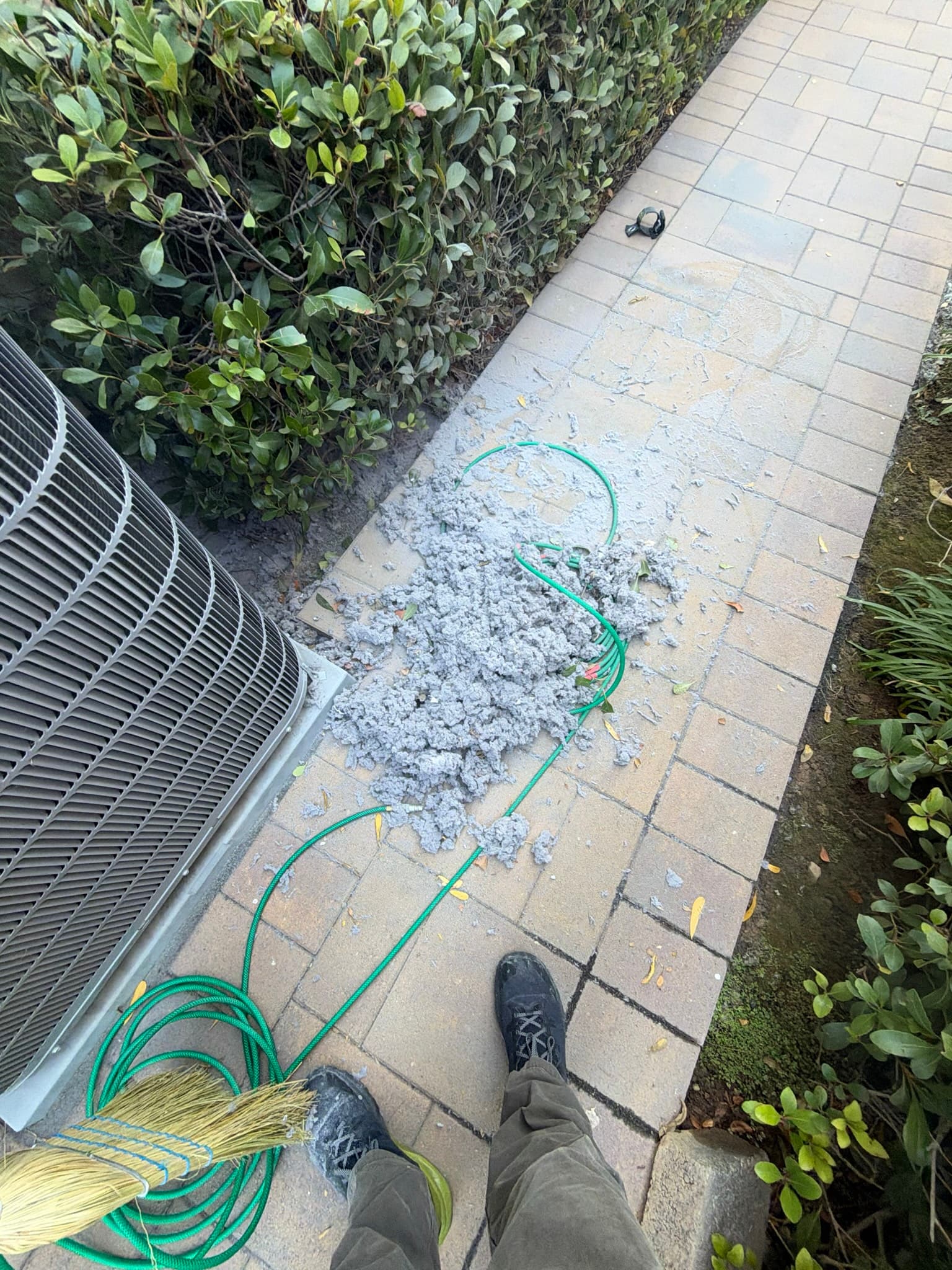 Large pile of extracted gray lint on pavers next to the rotary brush cable used to clean the dryer exhaust duct, with the condenser unit and hedge visible in the background.