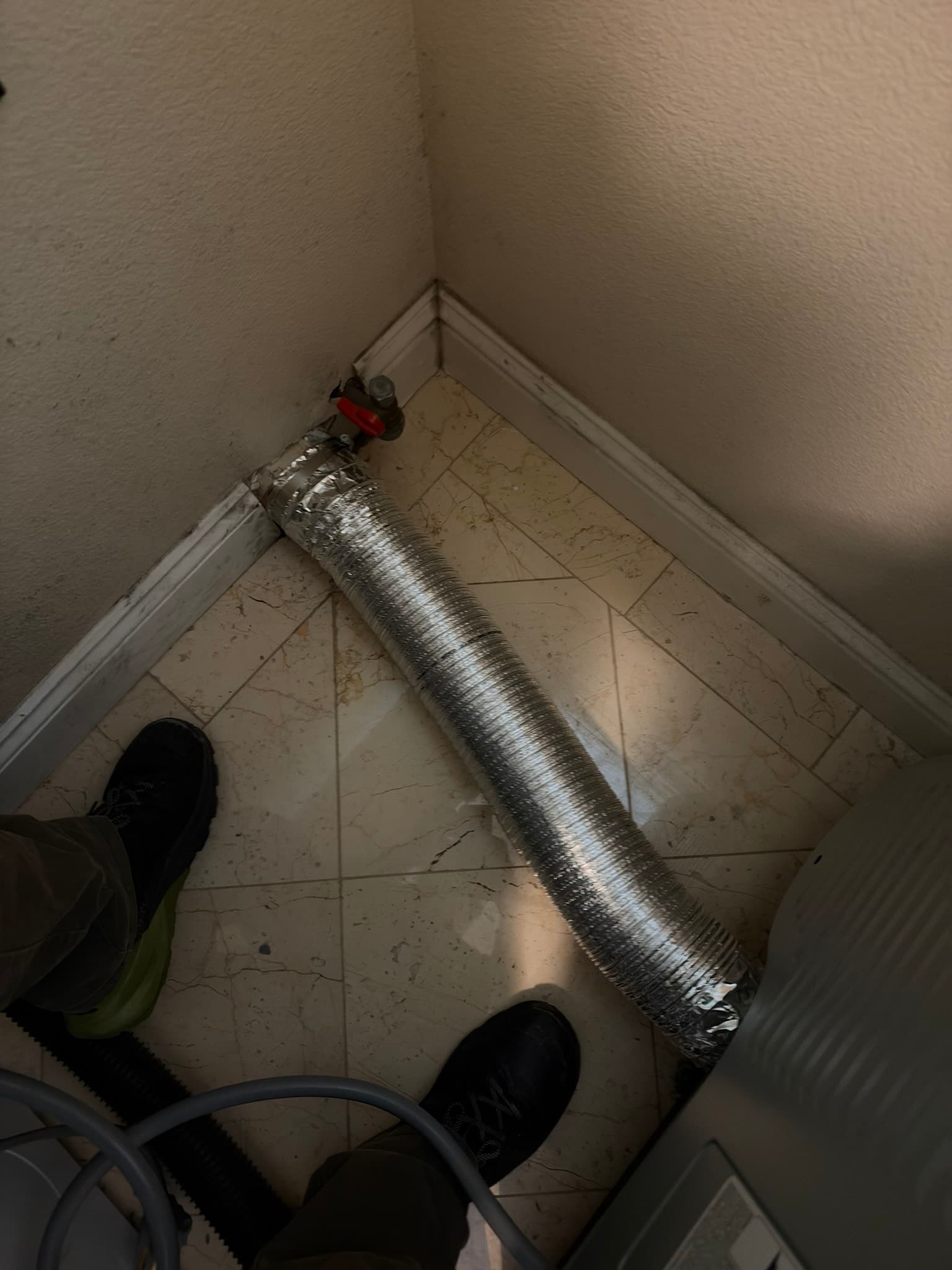 Flexible foil transition duct secured with foil tape at the wall connection point, running across a tile floor near a red-handled gas shutoff valve, with technician shoes visible for scale.