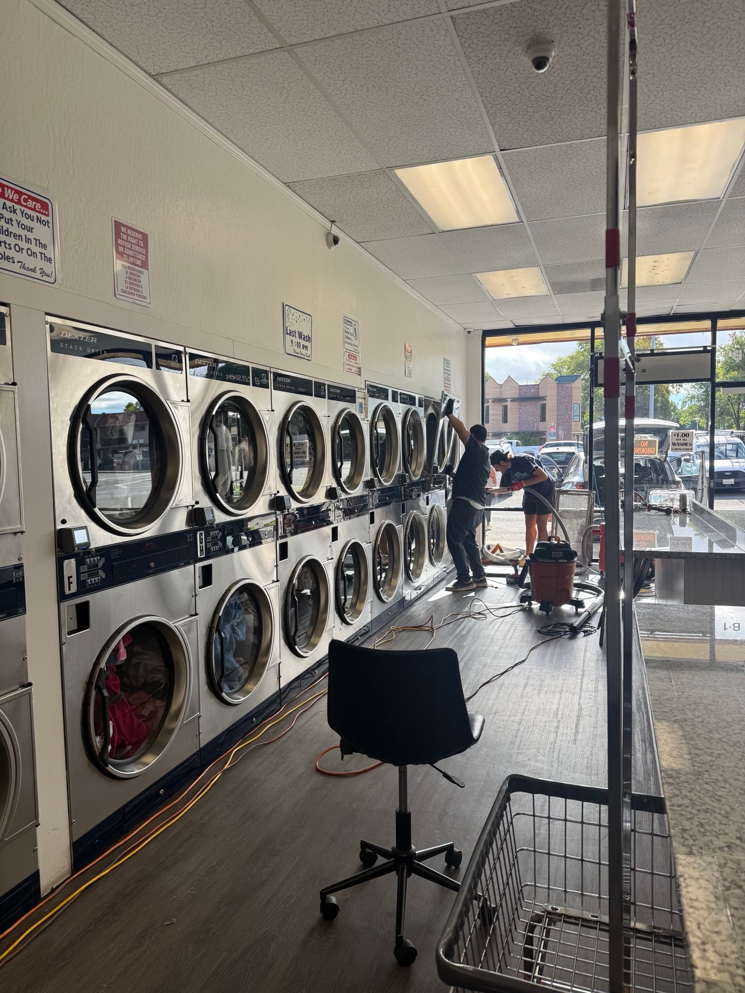 Wide-angle interior shot of the commercial laundromat showing stacked dryers mounted along the wall, a customer seating area in the foreground, and patrons using the facility