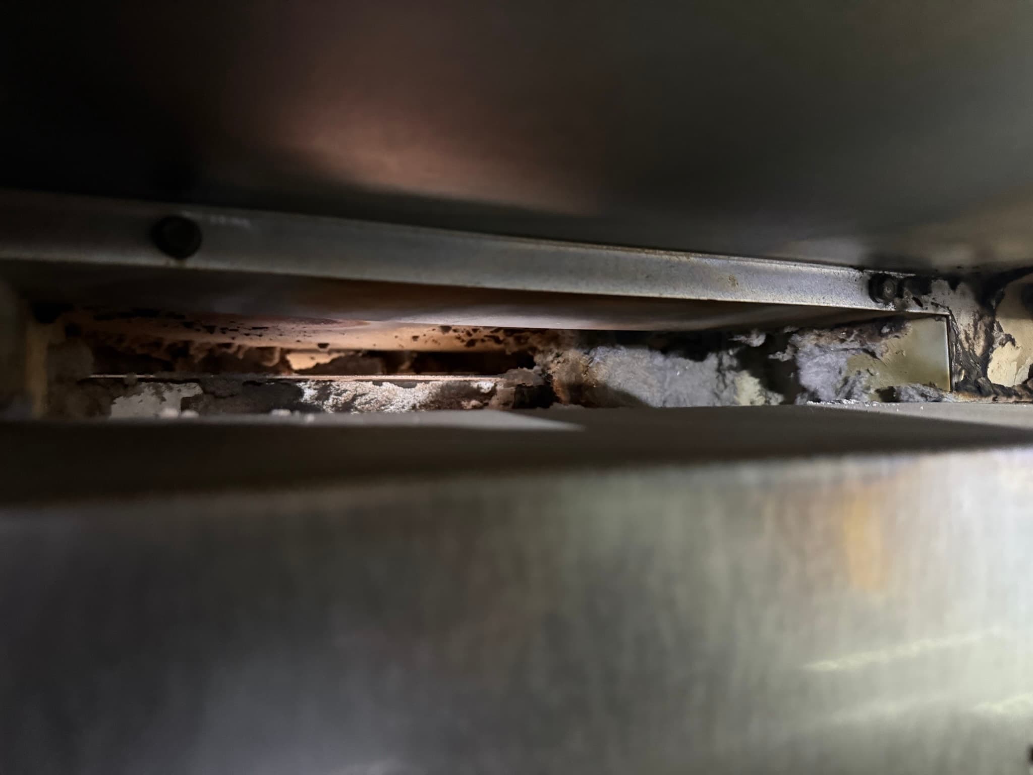 Close-up of a commercial dryer lint trap housing showing heavy compacted lint and dust accumulation mixed with discolored debris filling the trap cavity, indicating a long interval since last cleaning