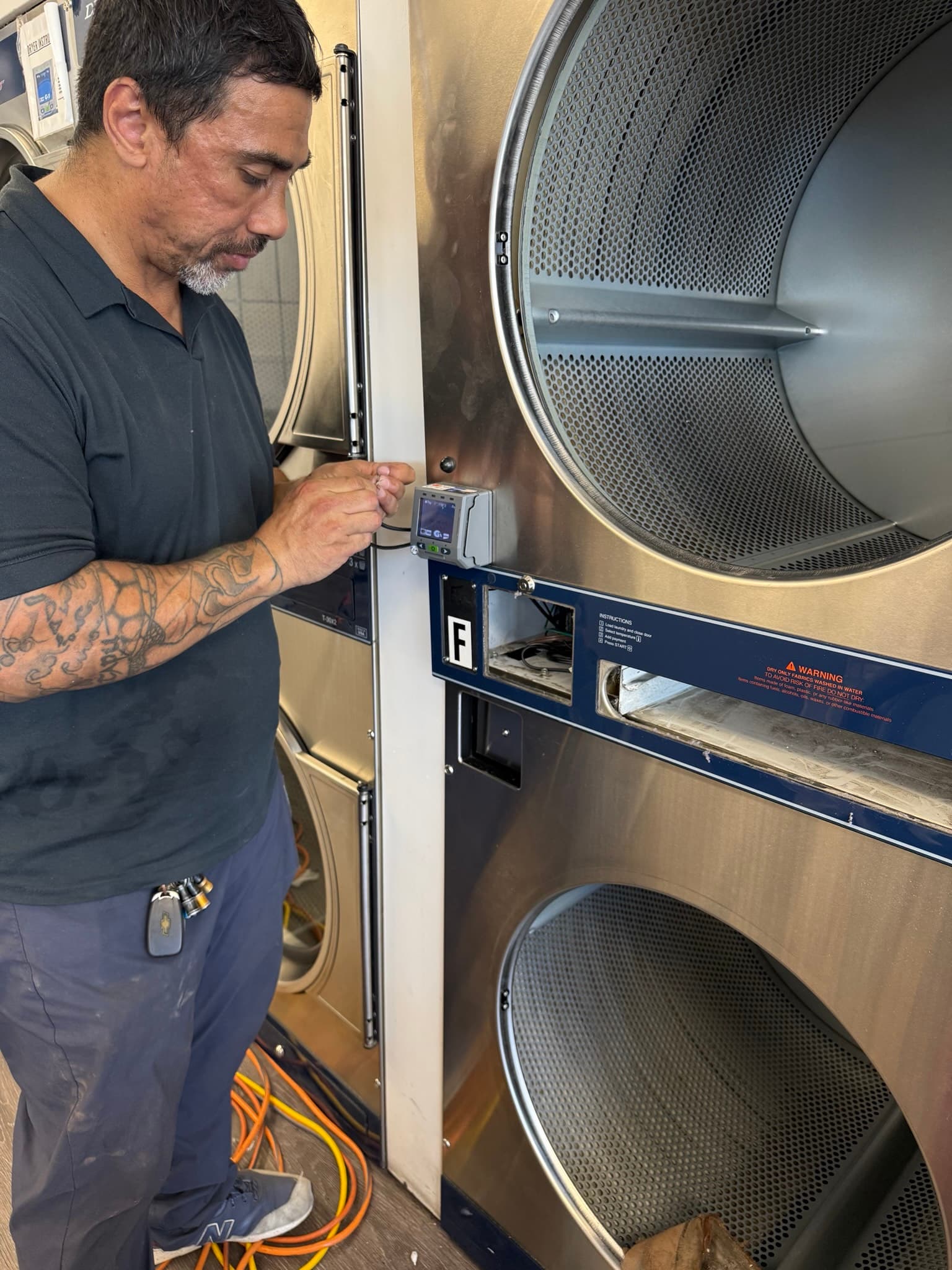 Technician holding a digital diagnostic meter near the control panel of an open commercial Dexter dryer, inspecting the clean perforated stainless steel drum interior during a service call at a San Leandro laundromat