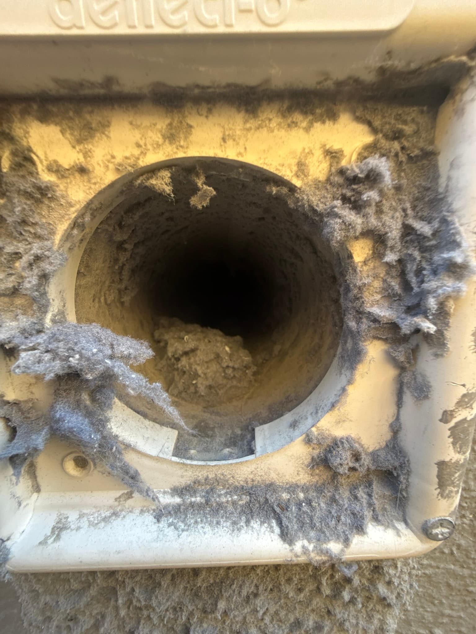 Close-up of a metal dryer exhaust duct opening packed with severe lint accumulation, partially blocking the duct interior and coating the surrounding mounting surface