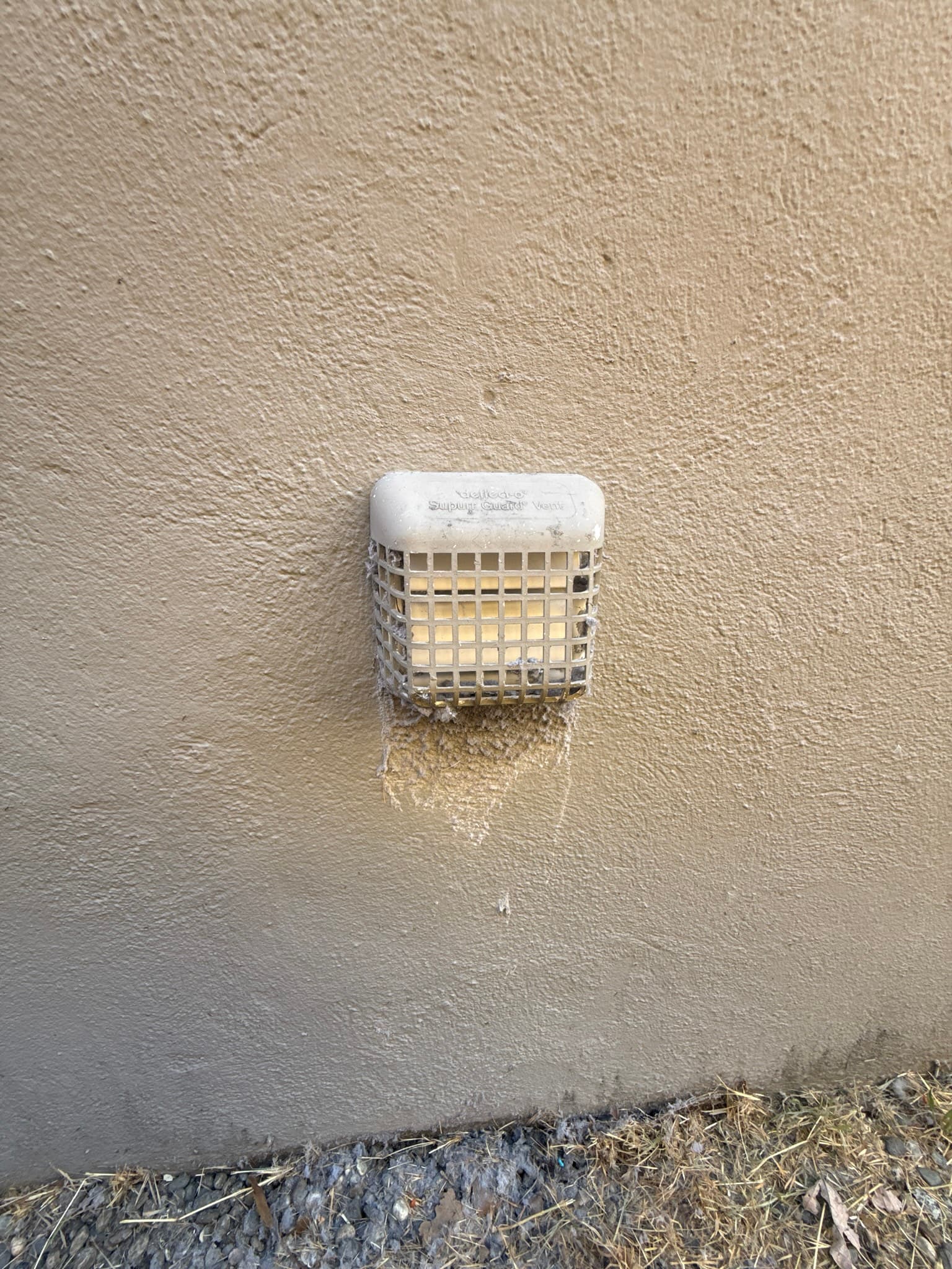 Exterior dryer vent cap with a plastic grid design mounted on a stucco wall, showing moisture staining and discoloration below the cap opening consistent with long-term condensation or restricted exhaust drainage.