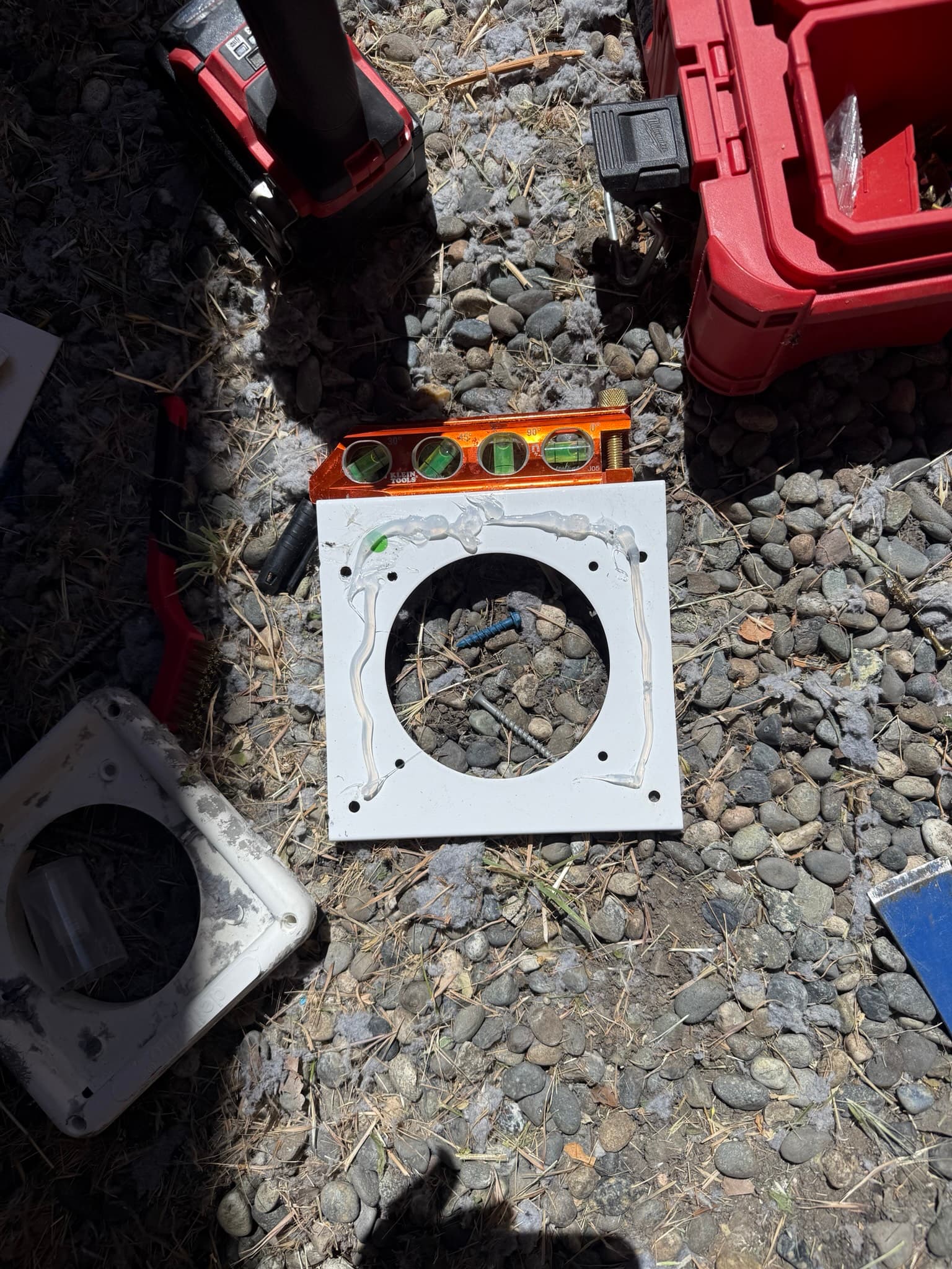 The new exterior wall vent mounting flange laid flat on gravel prior to installation, showing the central duct opening, perimeter mounting holes, and silicone caulk applied to the back surface for weatherproof sealing. An orange level tool is positioned above the component.