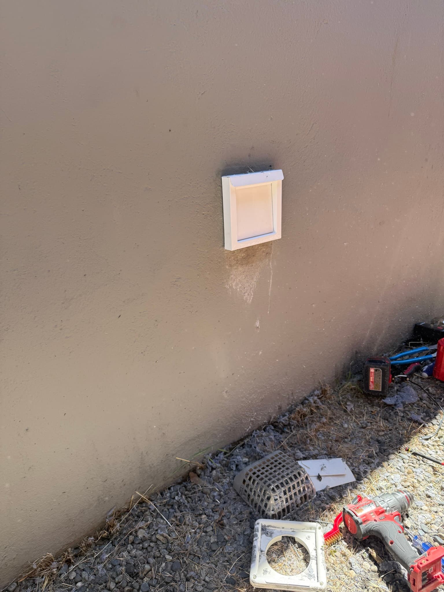 New white periscope-style dryer vent cap mounted on a concrete foundation wall after installation, with vent components and tools visible on the ground below