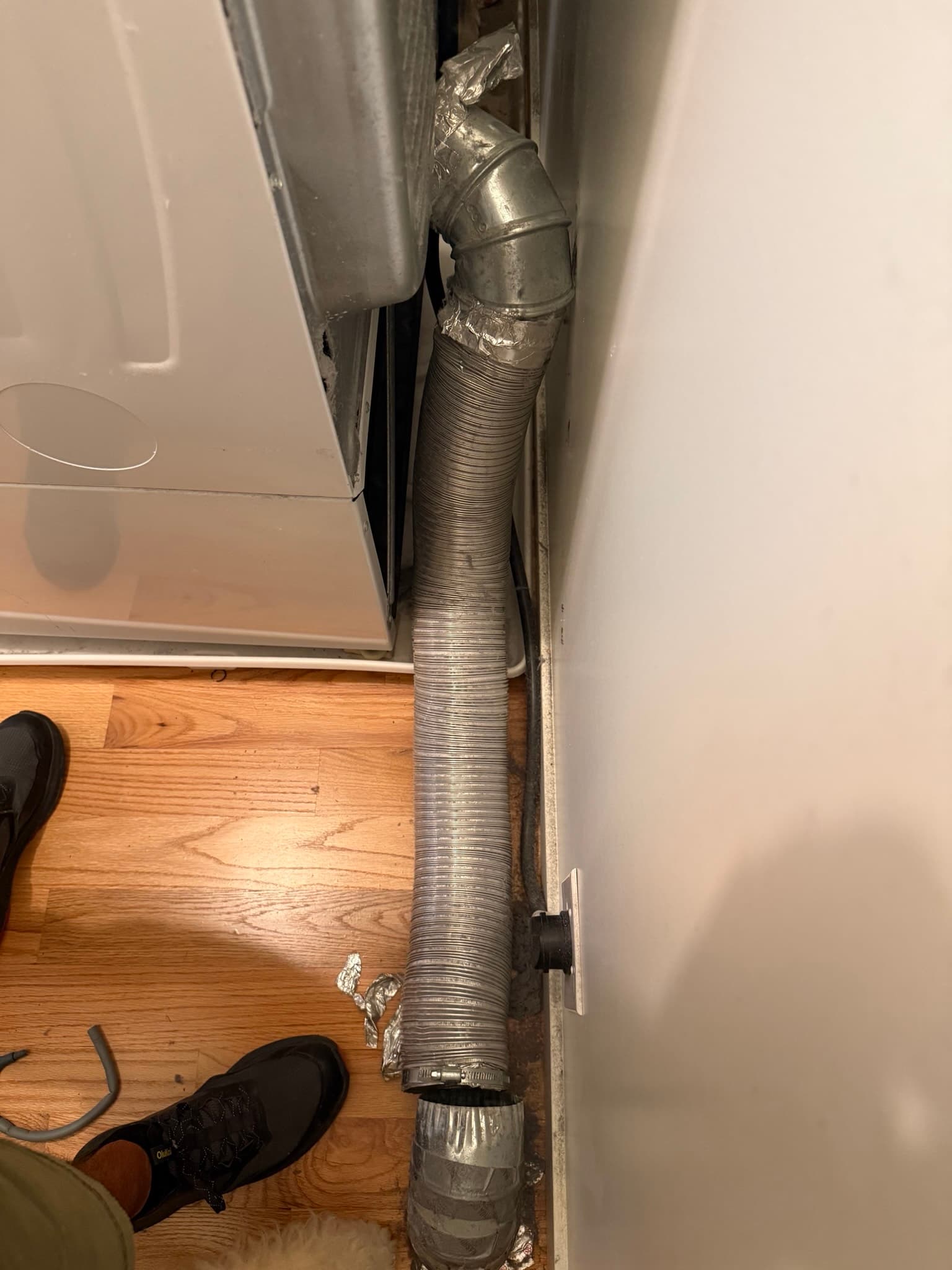 Flexible metal dryer vent duct with code violations: joints wrapped in cloth duct tape instead of approved clamps, duct running horizontally along the floor with visible exterior lint and dust buildup