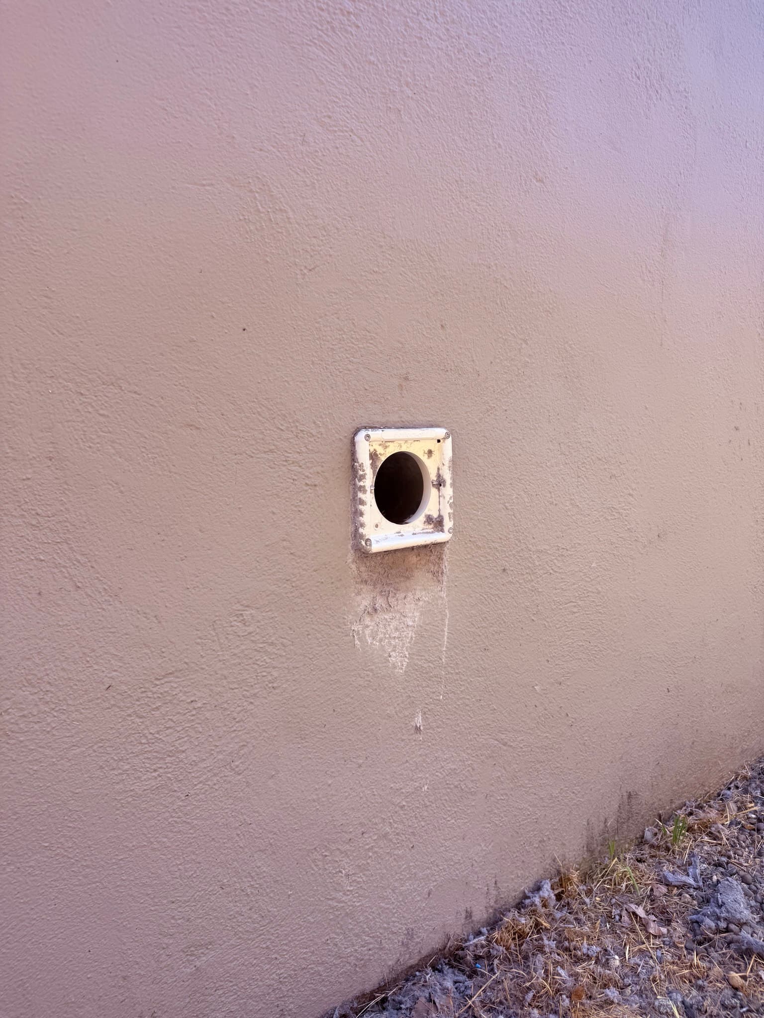Exterior dryer vent cap on a concrete or stucco wall showing dirty white metal mounting flange with discoloration and debris below the oval vent opening, installed near ground level