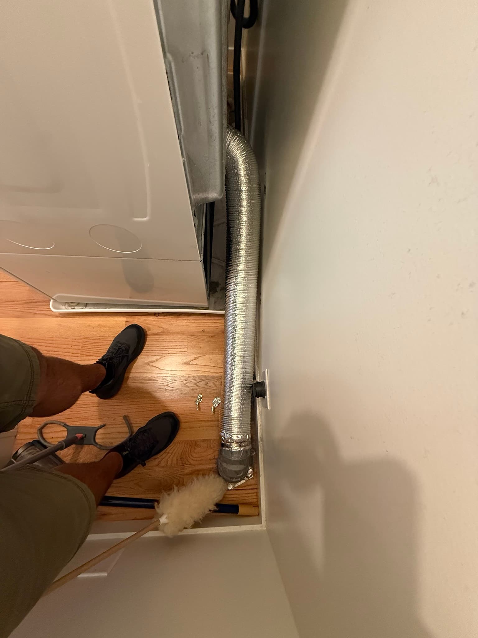 Rear of dryer unit showing a metallic flexible transition duct secured with reflective tape and metal clamps, with lint debris visible on the floor beneath the connection point