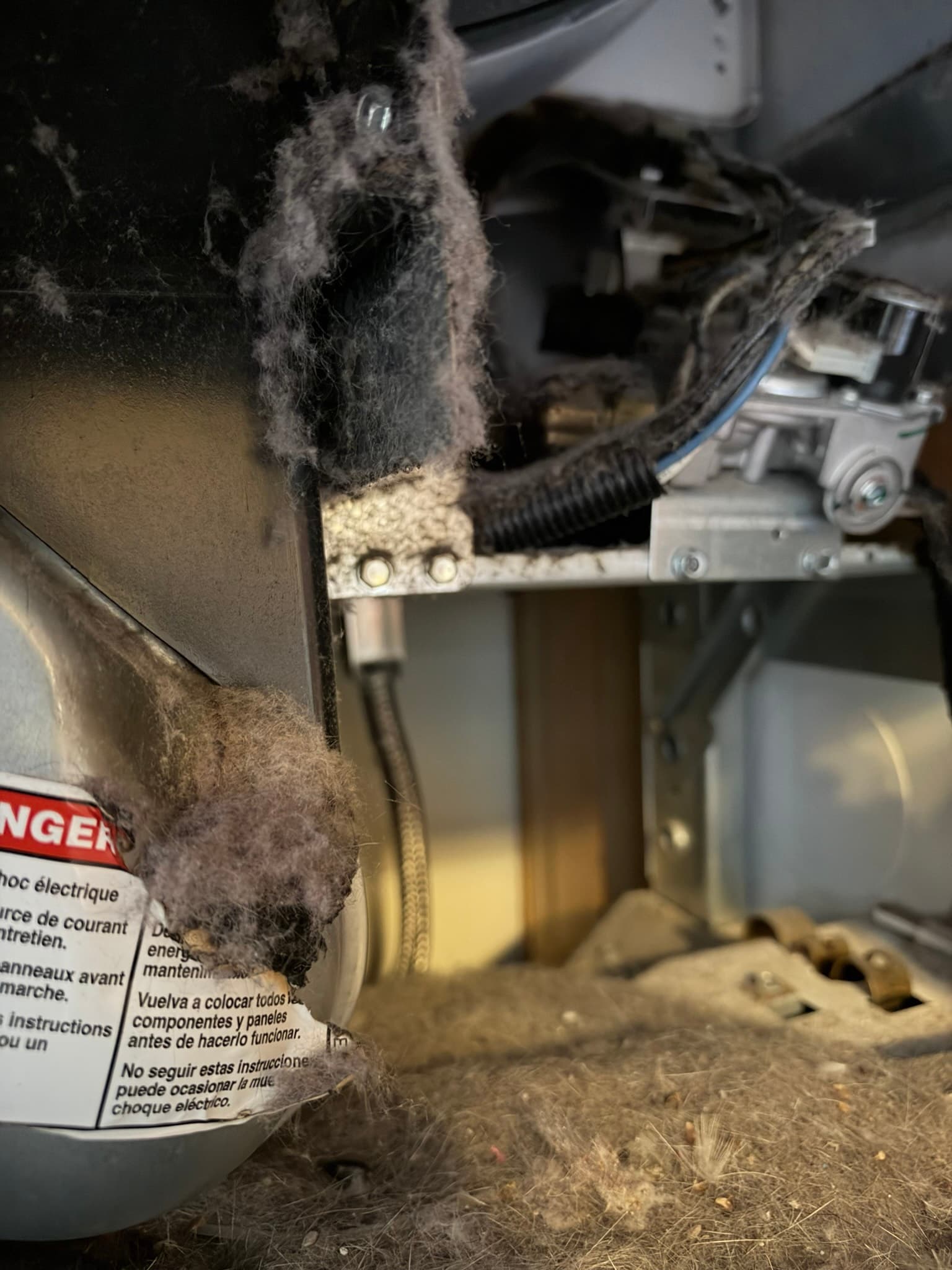 Dryer back panel removed to expose the drum, blower housing, and gas valve assembly matted with heavy lint accumulation across the burner compartment and wiring