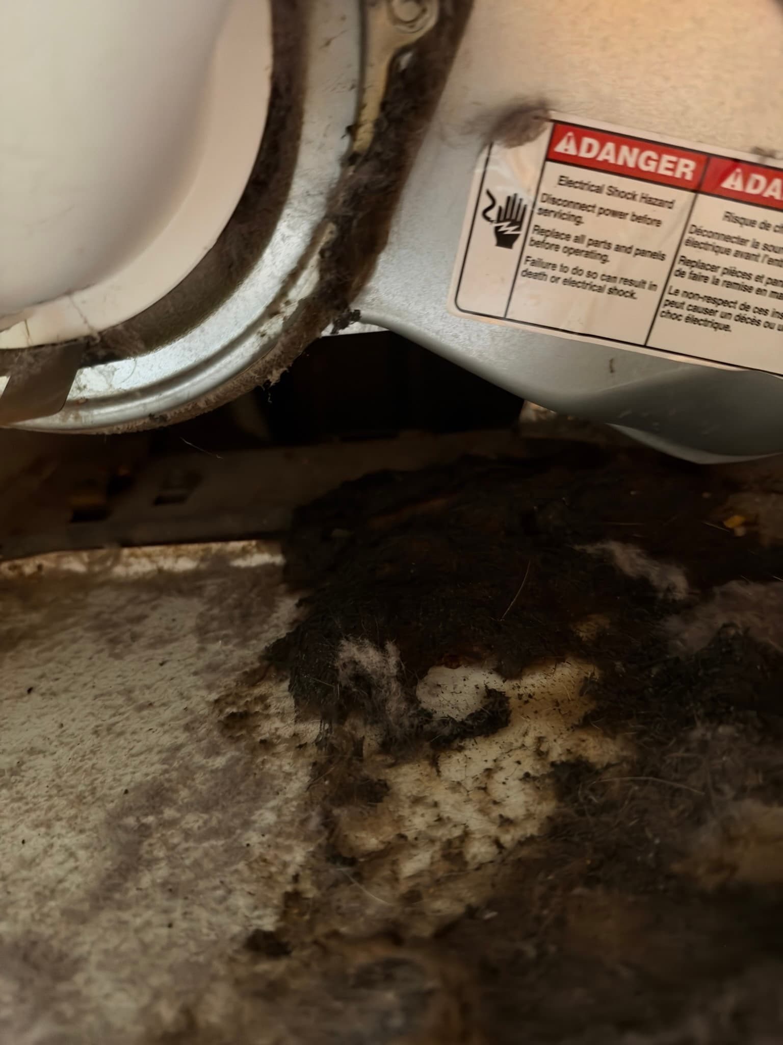 Dryer back panel exhaust outlet with the duct connection pulled back, showing heavy matted lint packed around the opening and spilled onto the floor beneath the unit