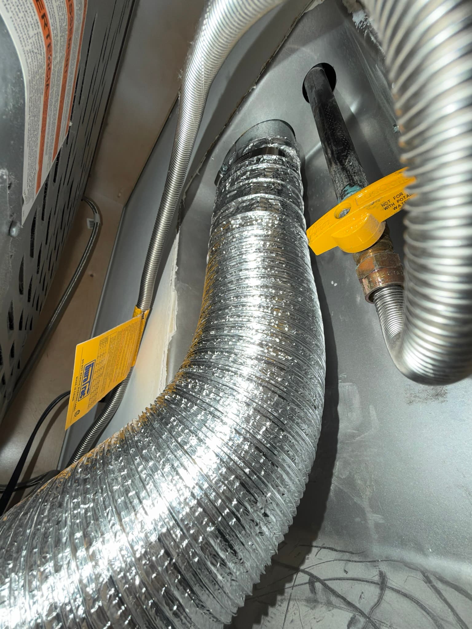 Rear of the dryer showing the foil flex transition duct clamped to the exhaust outlet alongside a gas supply line with a yellow shutoff valve