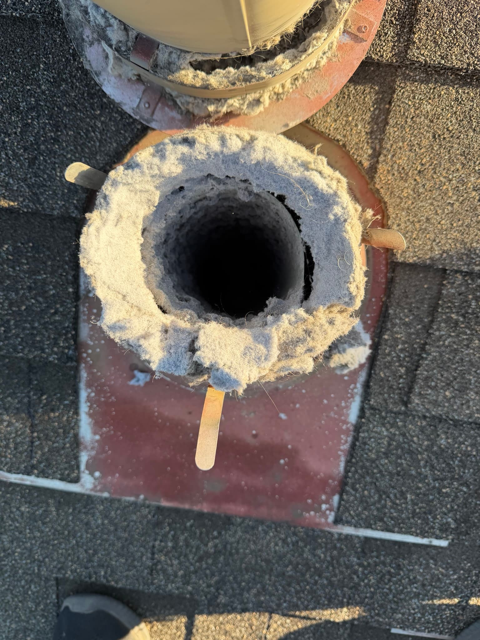 Overhead view of a metal roof dryer vent with lint matted around the opening and flashing, extending onto nearby shingles