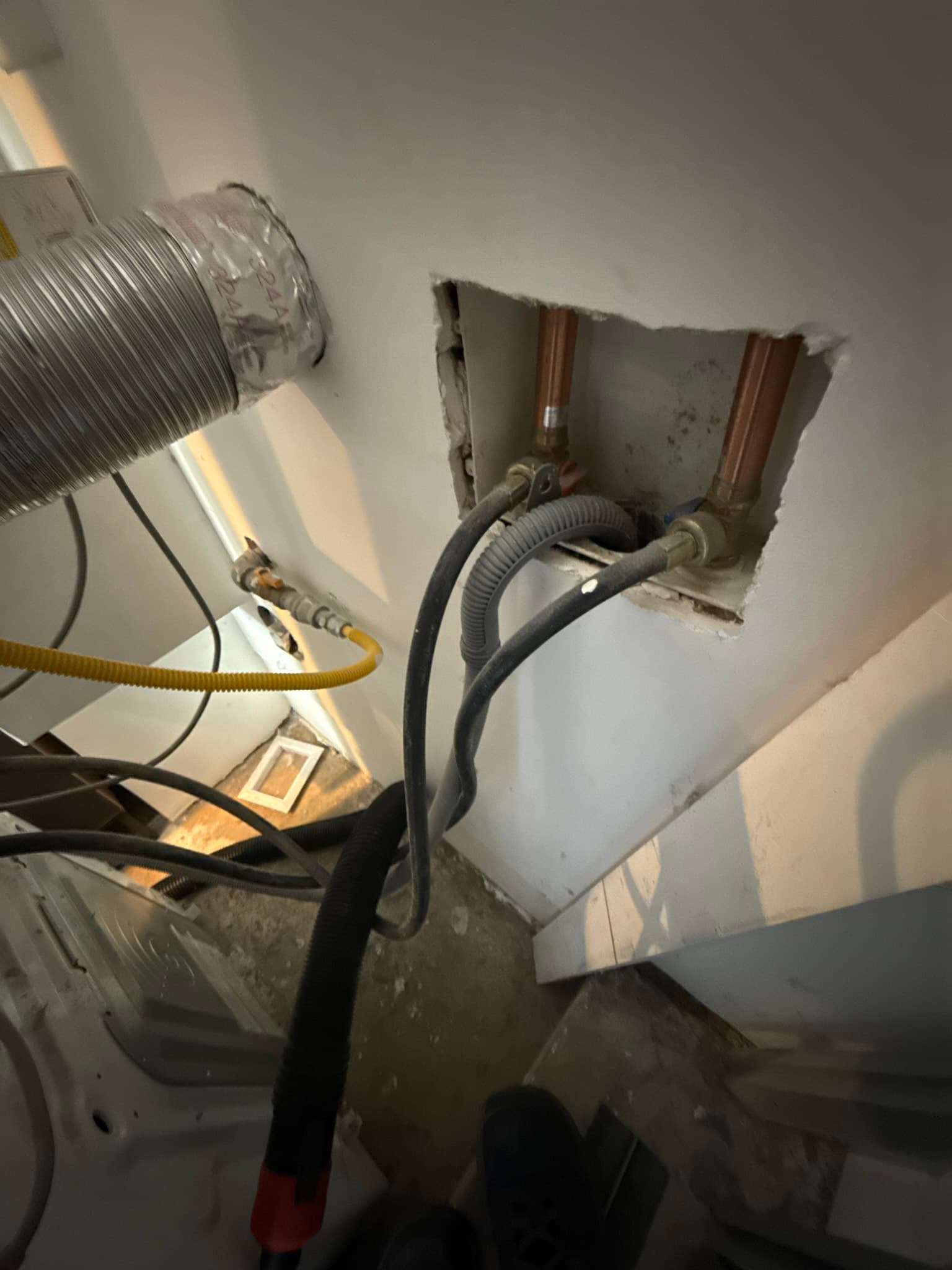 Dryer vent wall penetration area showing two flexible ducts entering a wall box with visible dust and debris accumulation around the connections and gaps visible around the ductwork