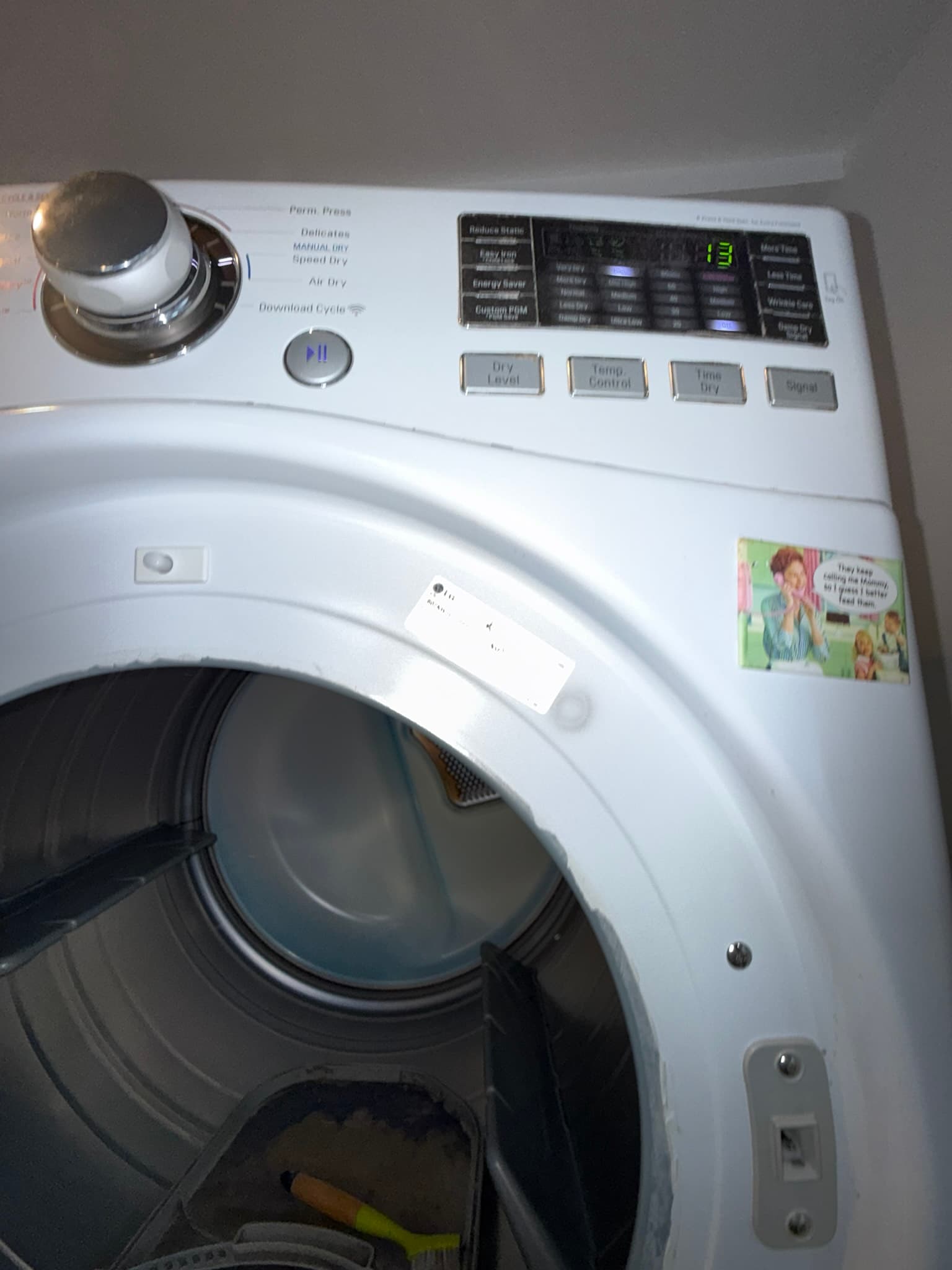 Front-loading dryer drum interior with the door open, showing a clean drum and the control panel above displaying a digital readout and cycle options including Perm Press, Delicates, Air Dry, and Download Cycle