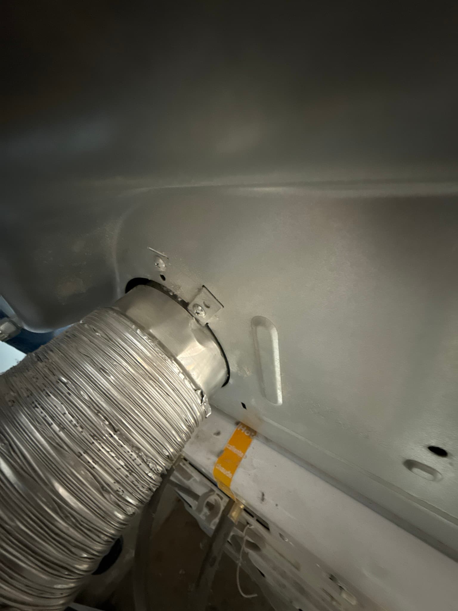 Rear of a clothes dryer showing a corrugated flexible aluminum transition duct connected to the dryer outlet with a metal band clamp, with a yellow identification label visible on the dryer cabinet