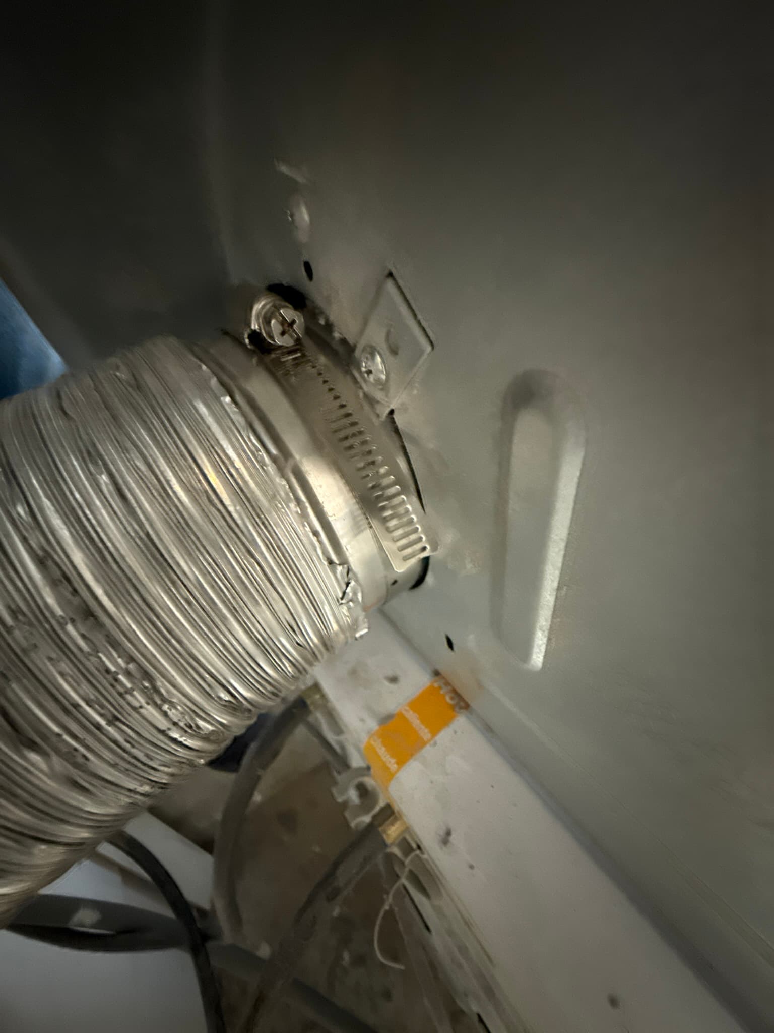 Flexible white dryer vent duct connected to a wall-mounted vent adapter with a worm-gear hose clamp, showing the wall connection point with a yellow label band visible