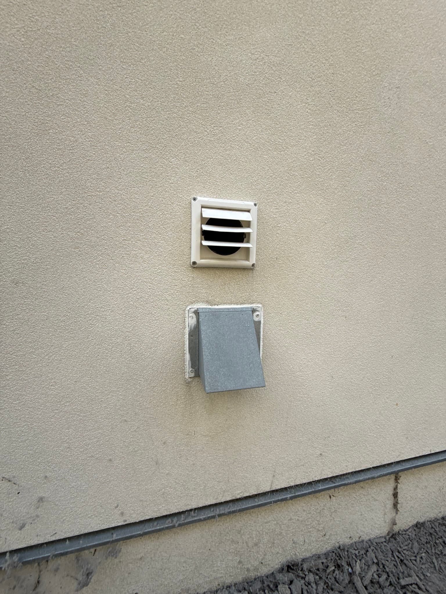 Two exterior dryer vent caps mounted on a stucco wall: the upper cap features horizontal louvered slats and appears clean, while the lower cap shows a gray damper flap in a closed position