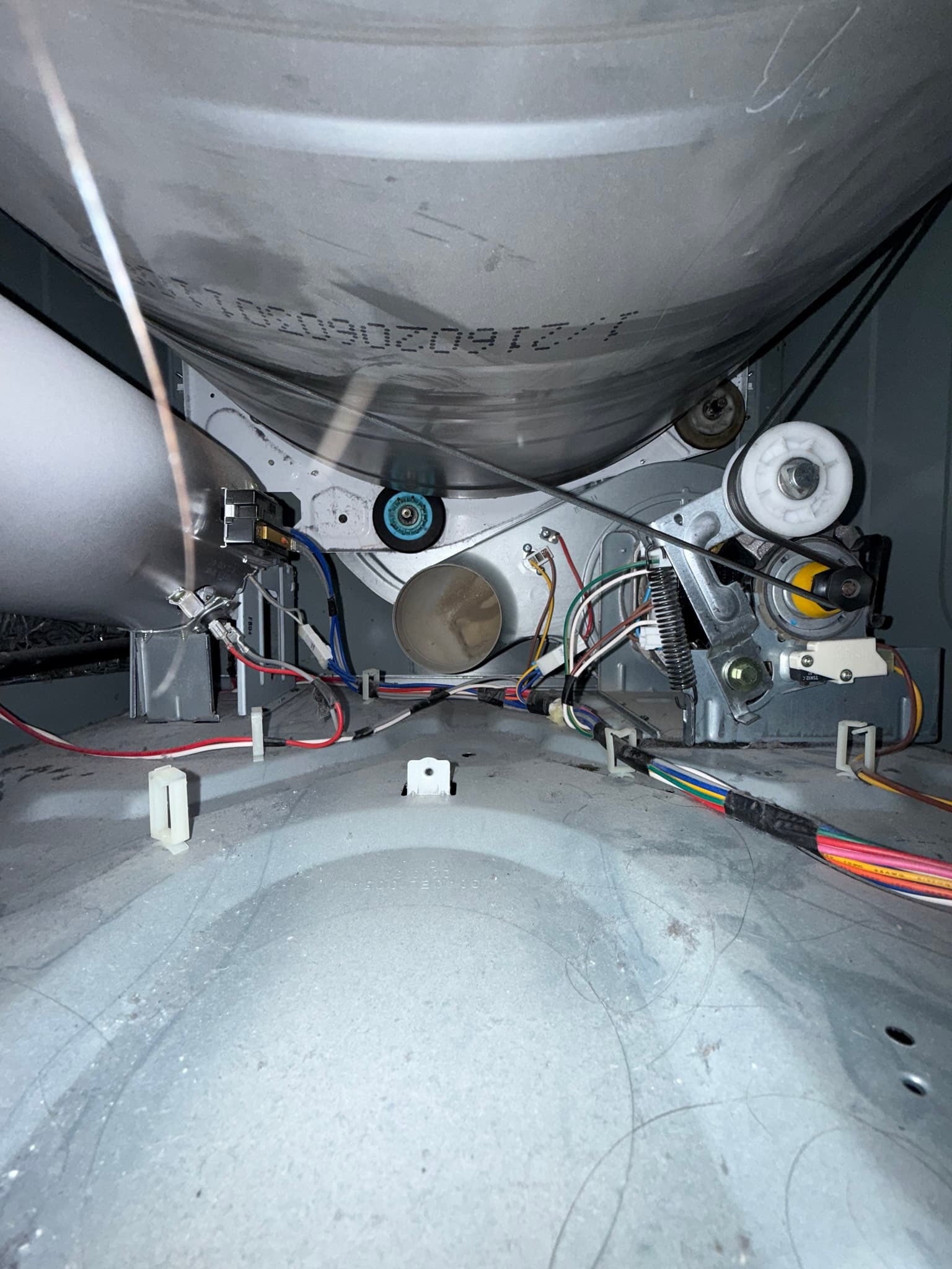 Interior underside of a clothes dryer drum showing light lint and dust accumulation on the metal surface, with mechanical components including the motor assembly and multicolored electrical wiring visible below