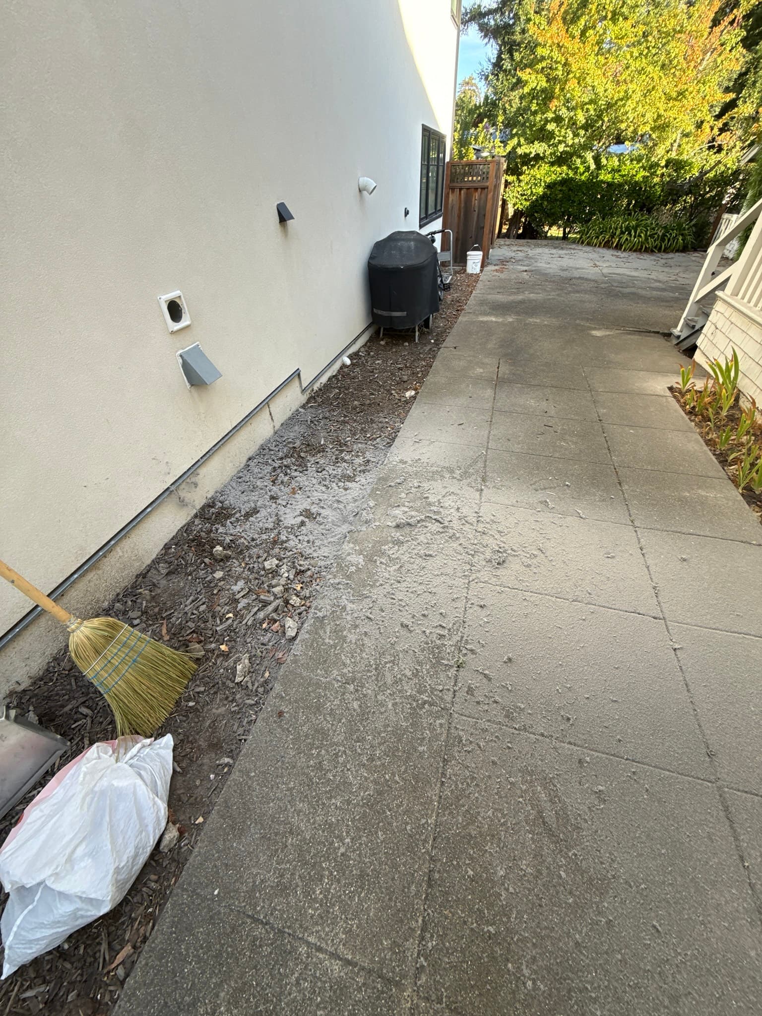 Exterior side wall of a residential structure with two white vent caps and a gray dryer vent cap visible, a narrow gravel strip along the foundation, and a broom and trash bag in the foreground