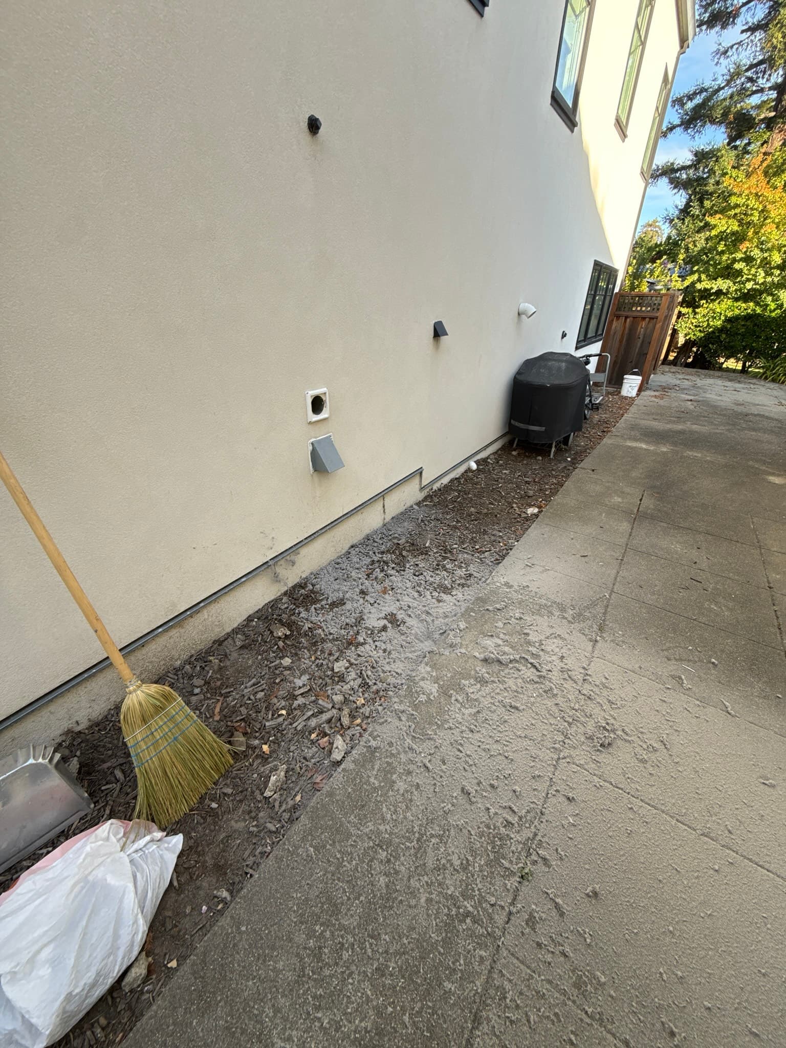 Exterior stucco wall of a residential building showing multiple white periscope-style dryer vent caps and black duct outlets at varying heights, with a broom and trash bags on the ground below indicating active cleanup